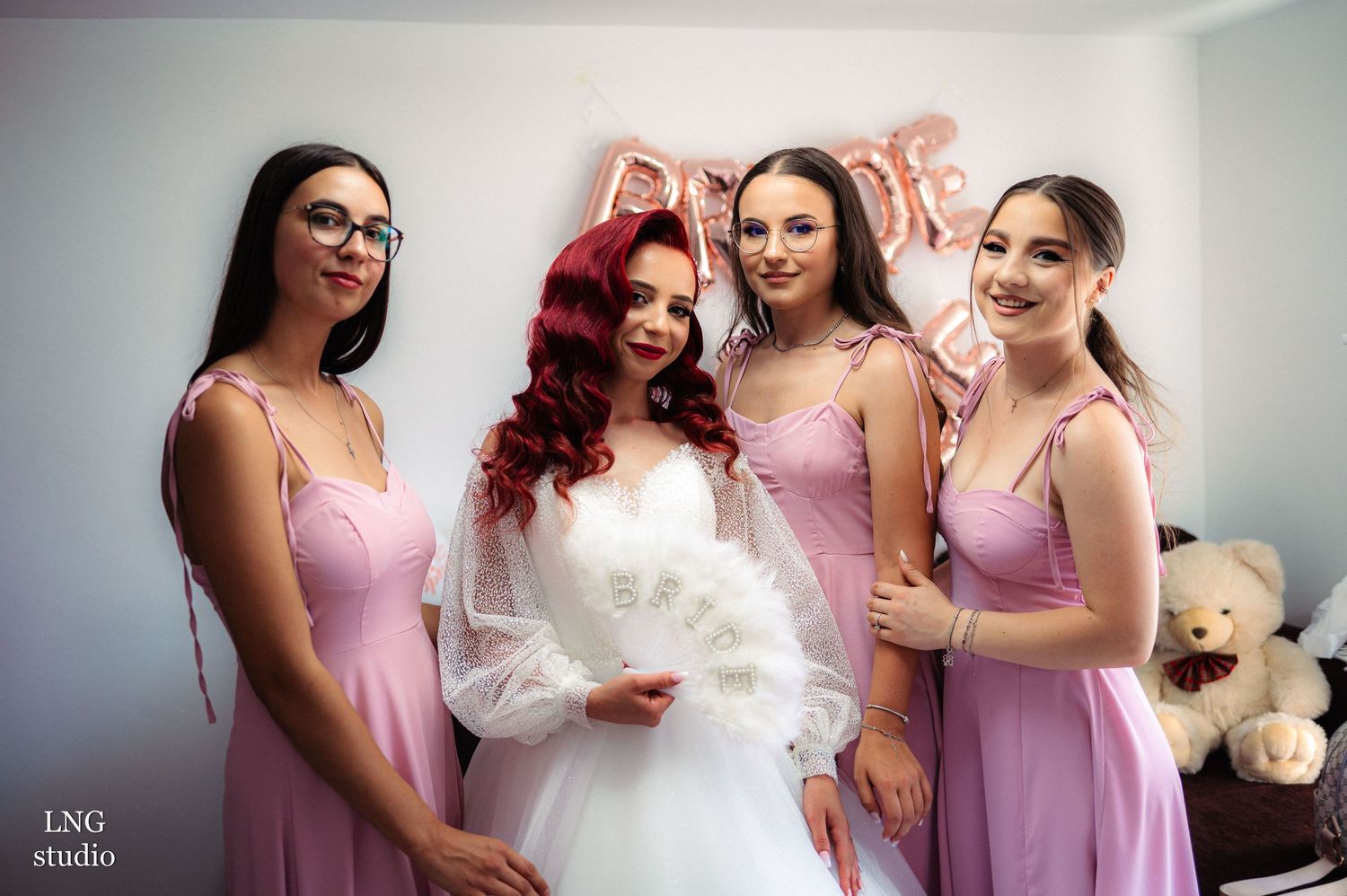 Wedding party in pink dresses poses together with bride under rose gold balloon decorations.