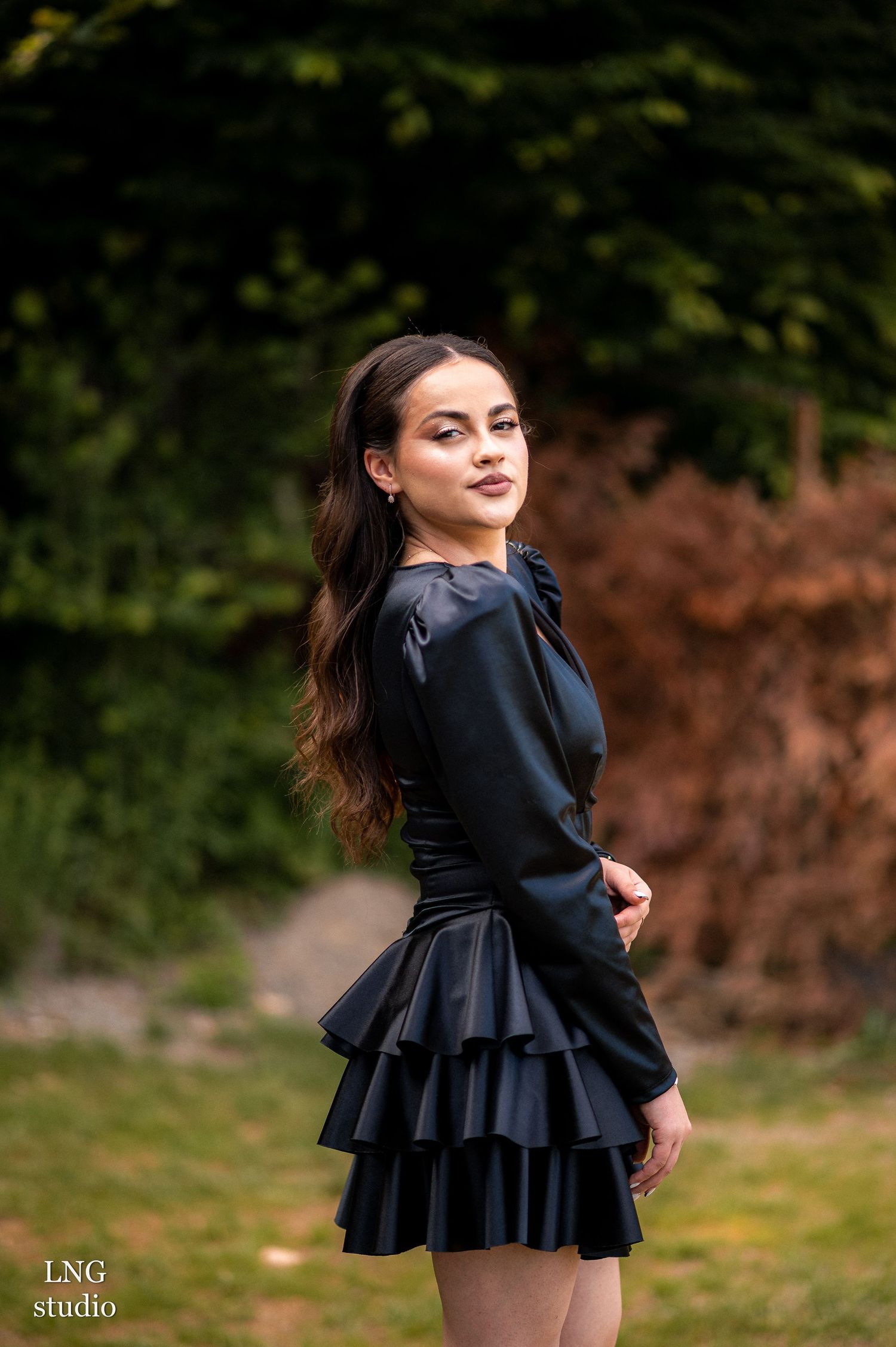 An outdoor portrait in a black long-sleeved dress with tiered ruffled skirt against a natural green background.