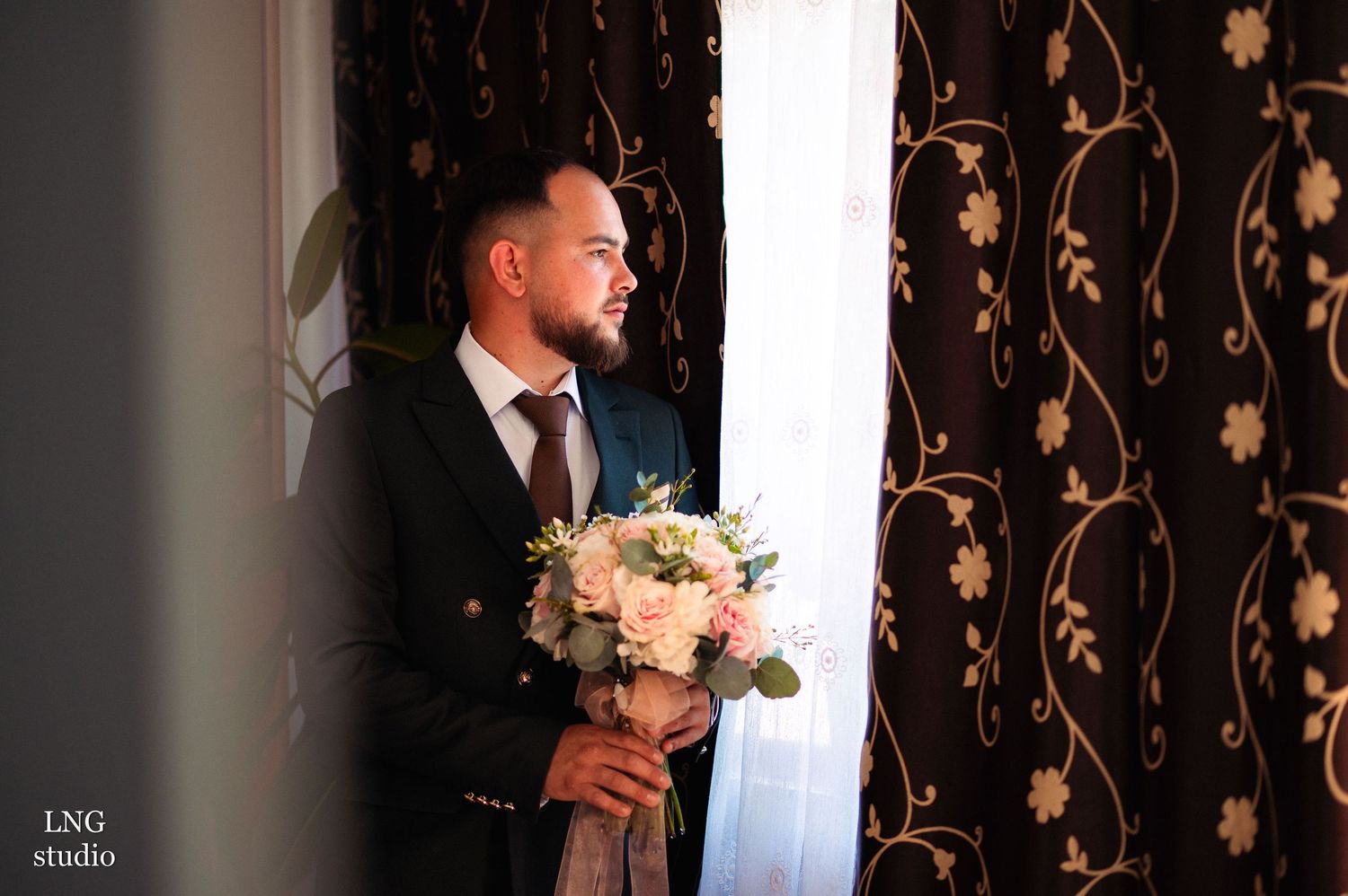 Groom holding elegant white floral bouquet against dark patterned backdrop.