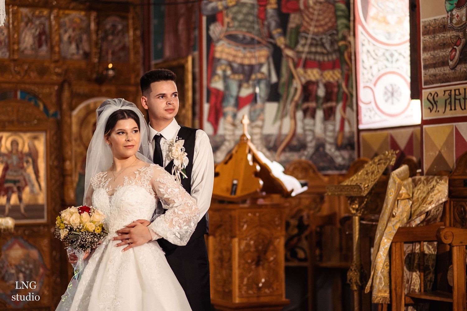 A wedding couple poses in an ornate church interior with traditional decorations and wooden furnishings.