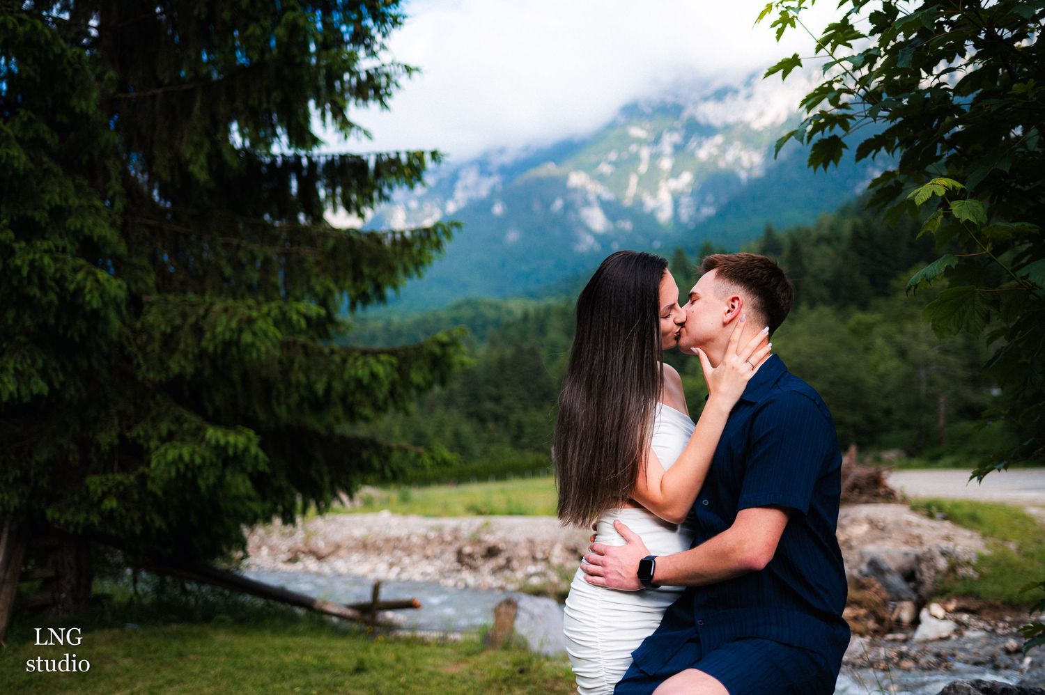 A couple shares a kiss with majestic mountains and evergreen trees in the background.