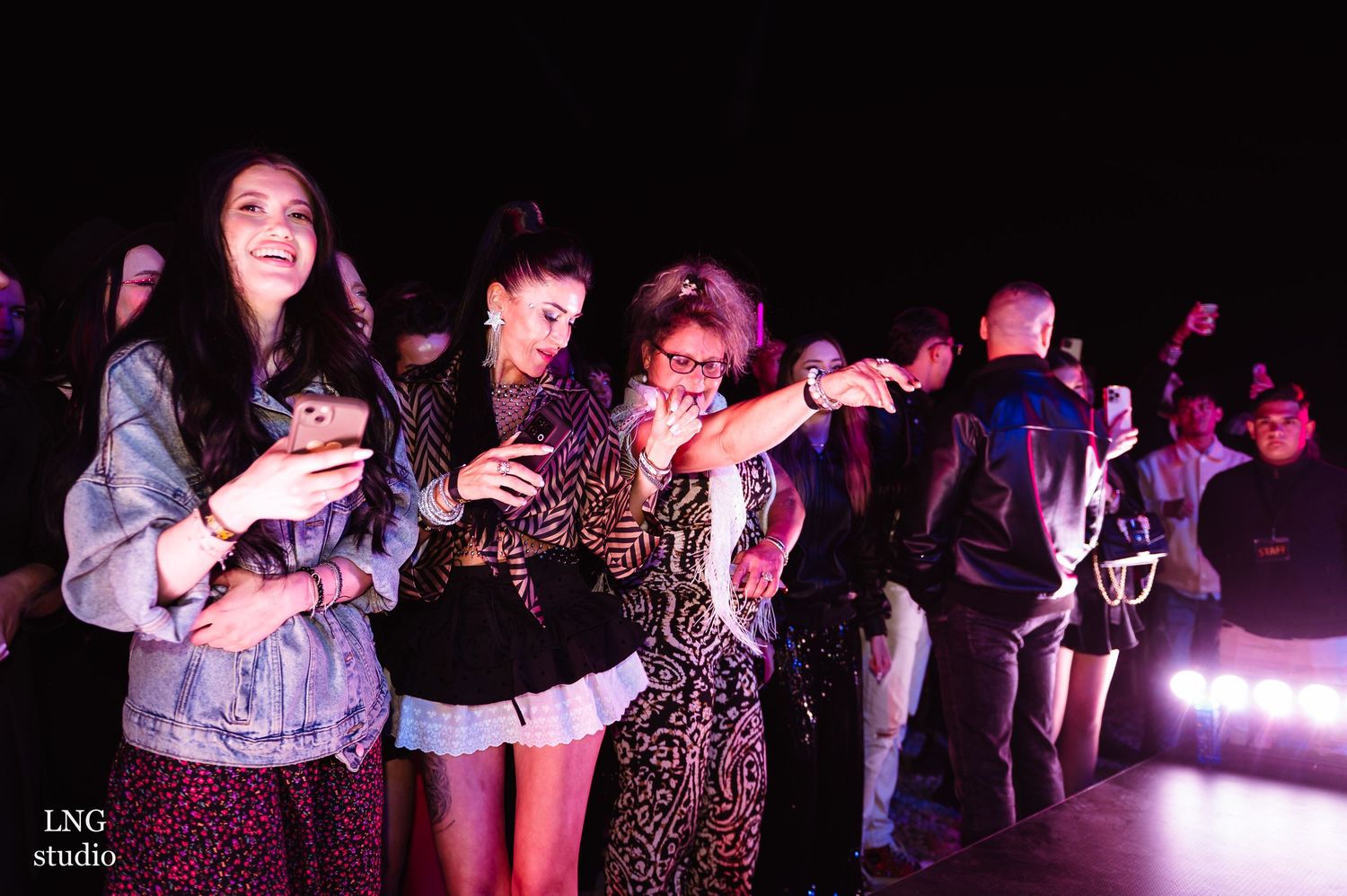 Concert attendees dance and smile while enjoying nighttime music performance with purple lighting.