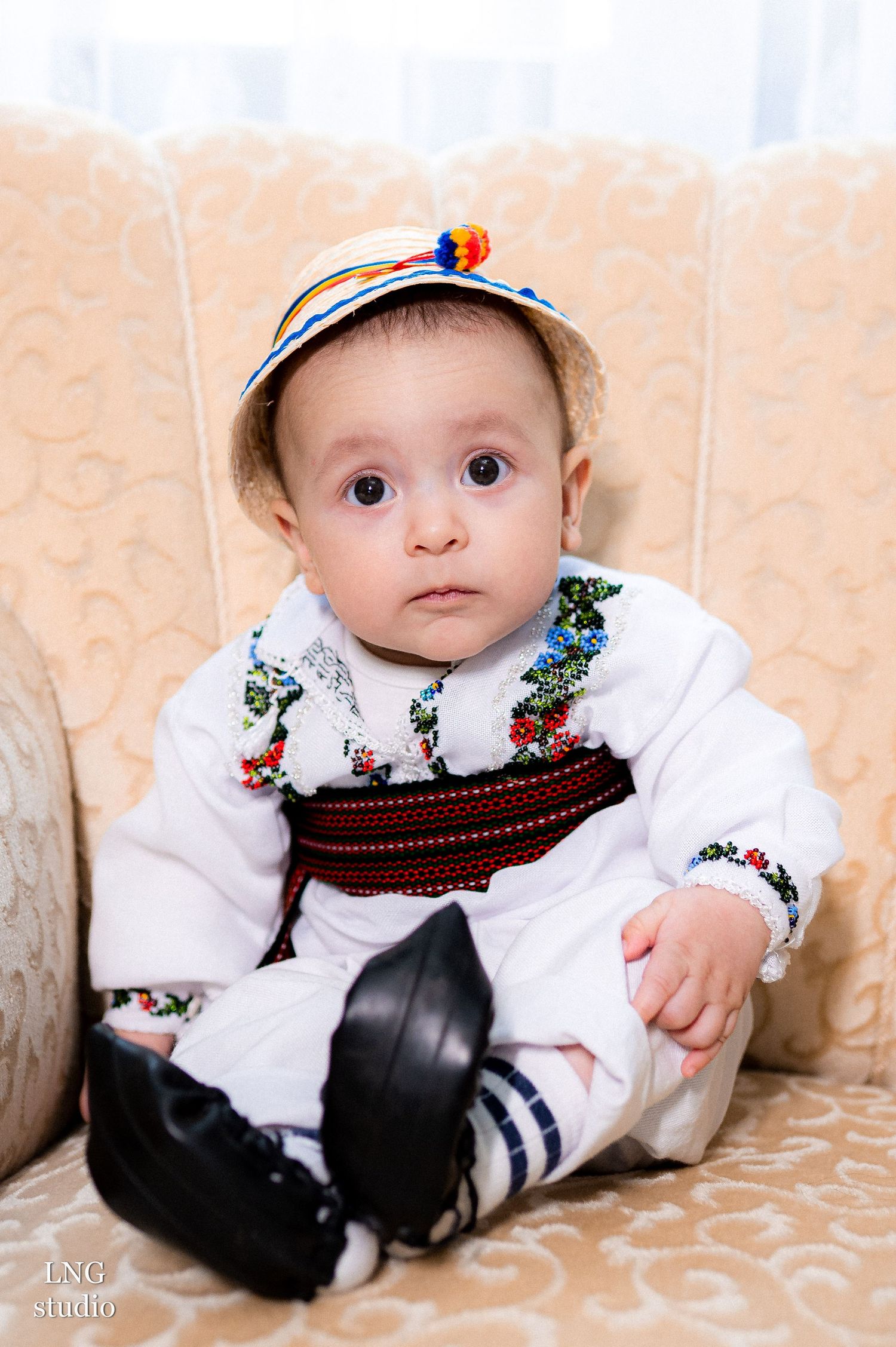 Baby wearing traditional Ukrainian folk costume with embroidery and decorative cap on beige couch.
