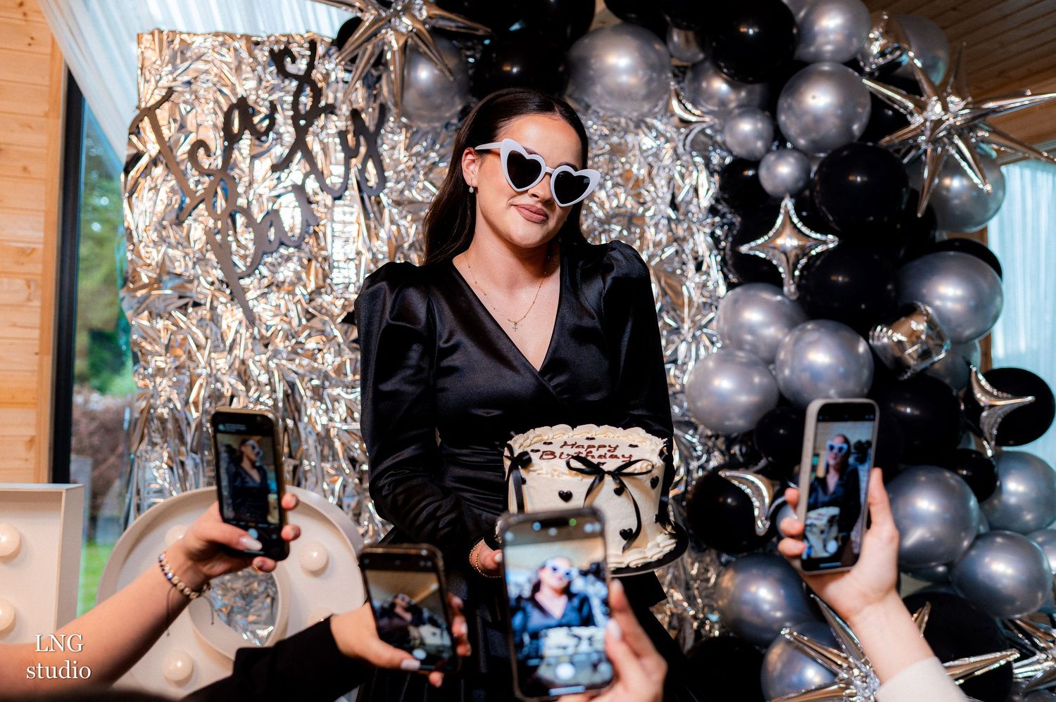 A person in black attire holding a gift box while others take photos against a silver and black balloon backdrop.