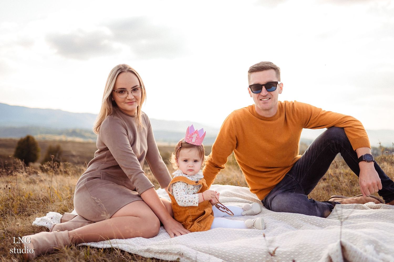 Family sits together on white blanket for first birthday photoshoot in rustic outdoor setting.