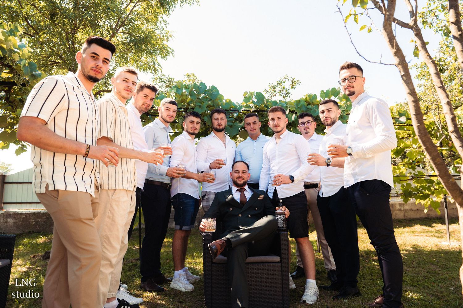 Groomsmen gather outdoors for casual group photo with drinks at a summer wedding celebration.