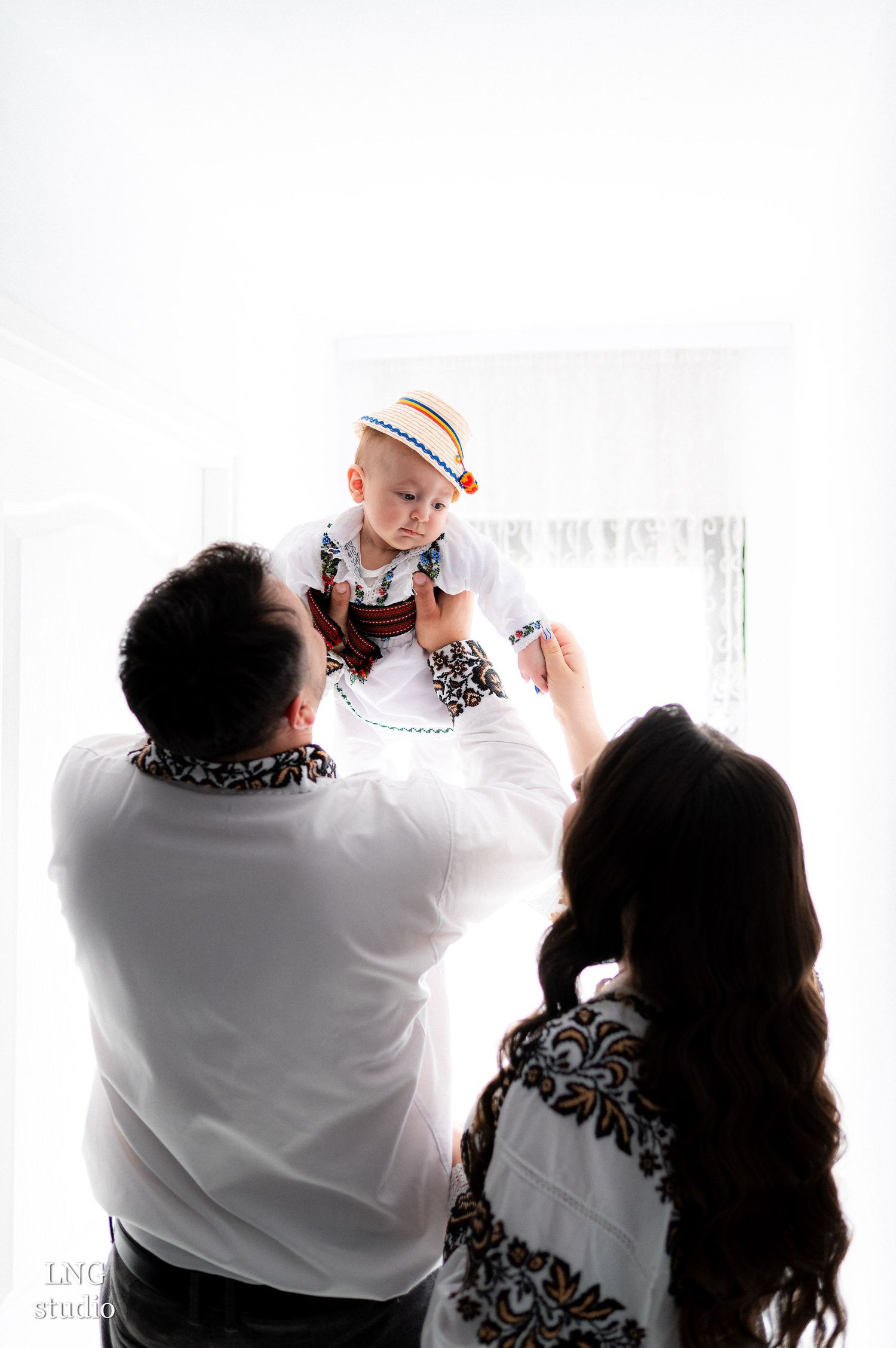 Family in traditional embroidered white shirts lifting baby during indoor photoshoot.