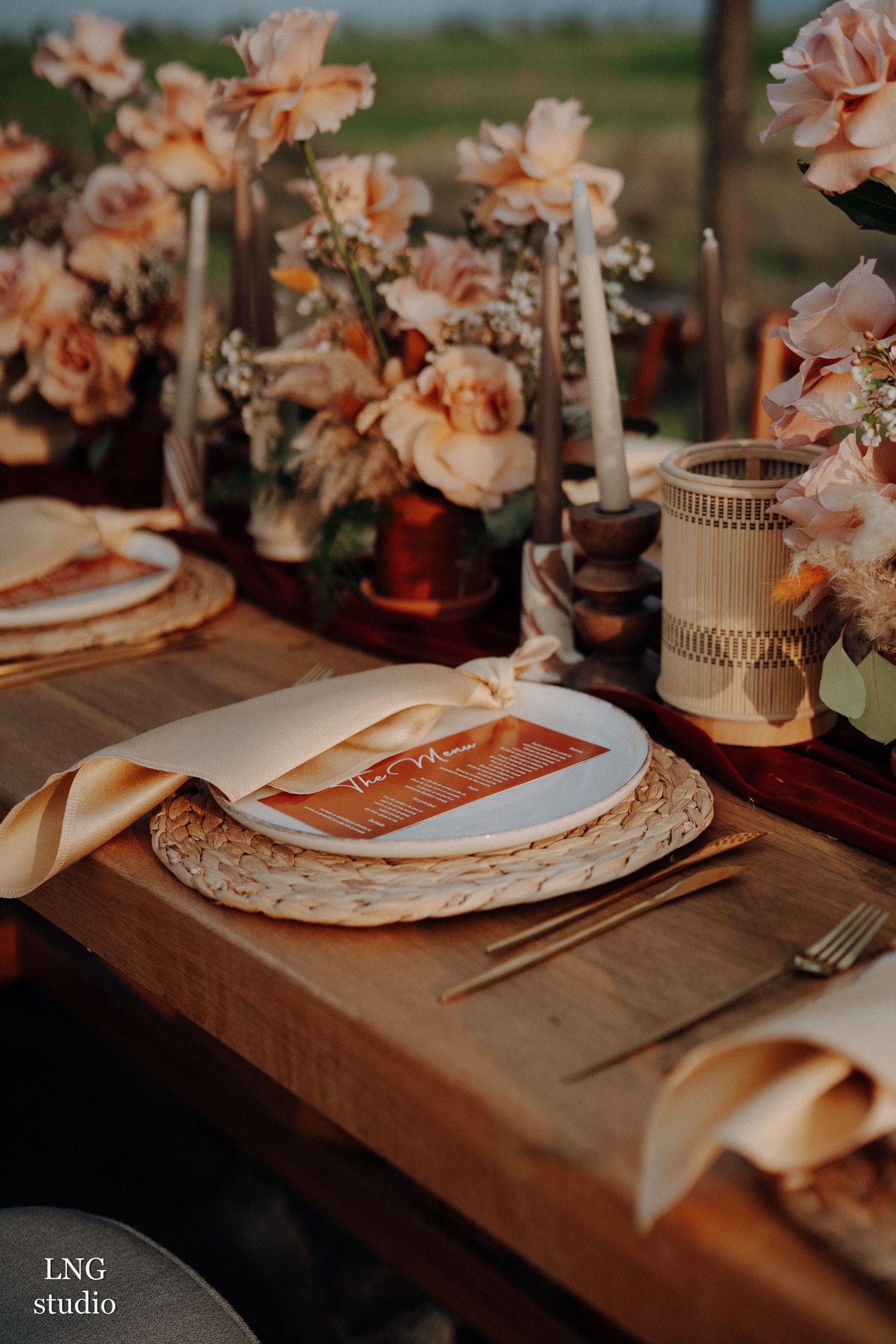 Elegant table setting with peach roses, taper candles, and woven charger plates on wooden surface in warm lighting.
