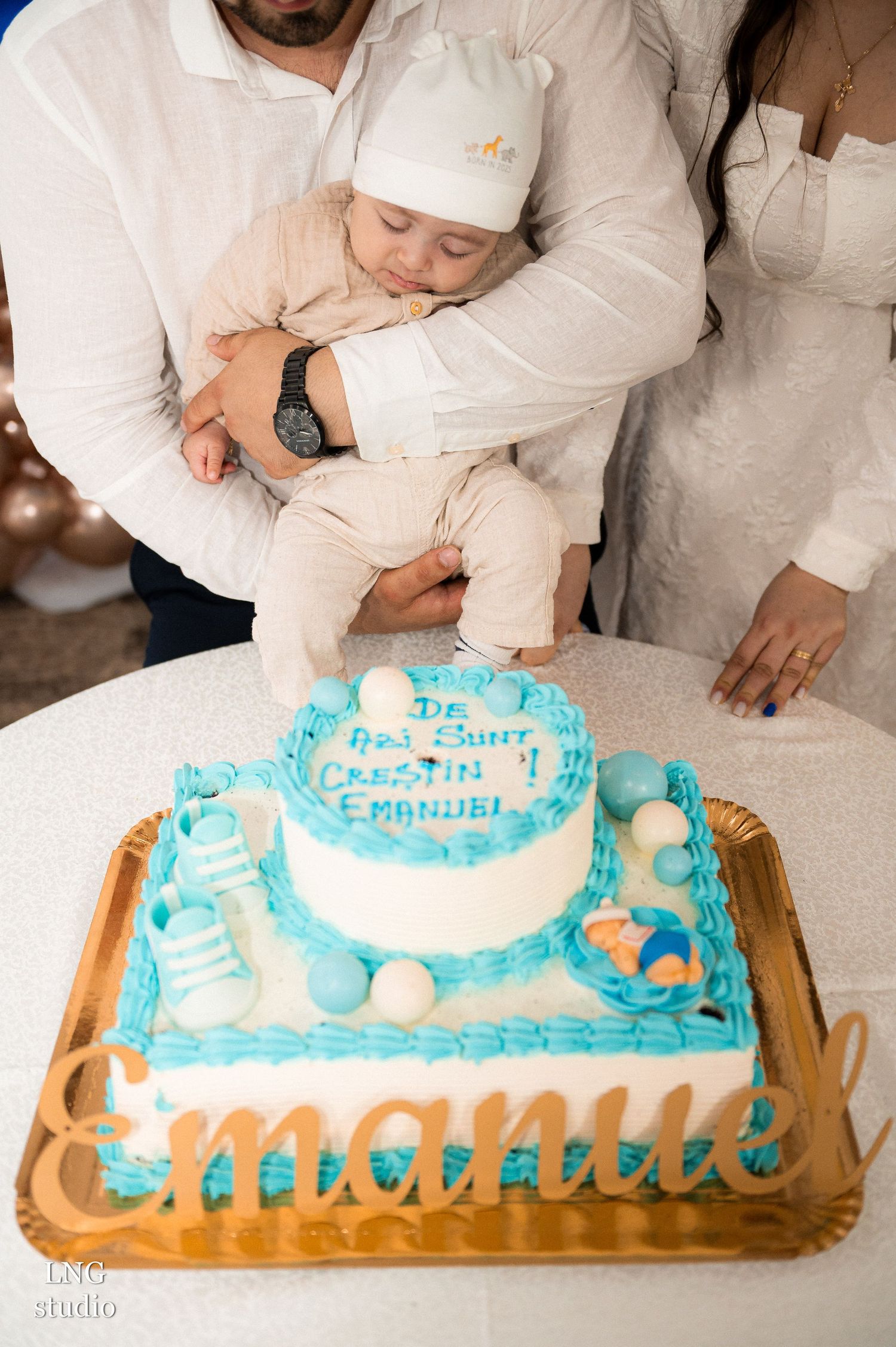 A baby in white clothing leans over a blue and white celebration cake.