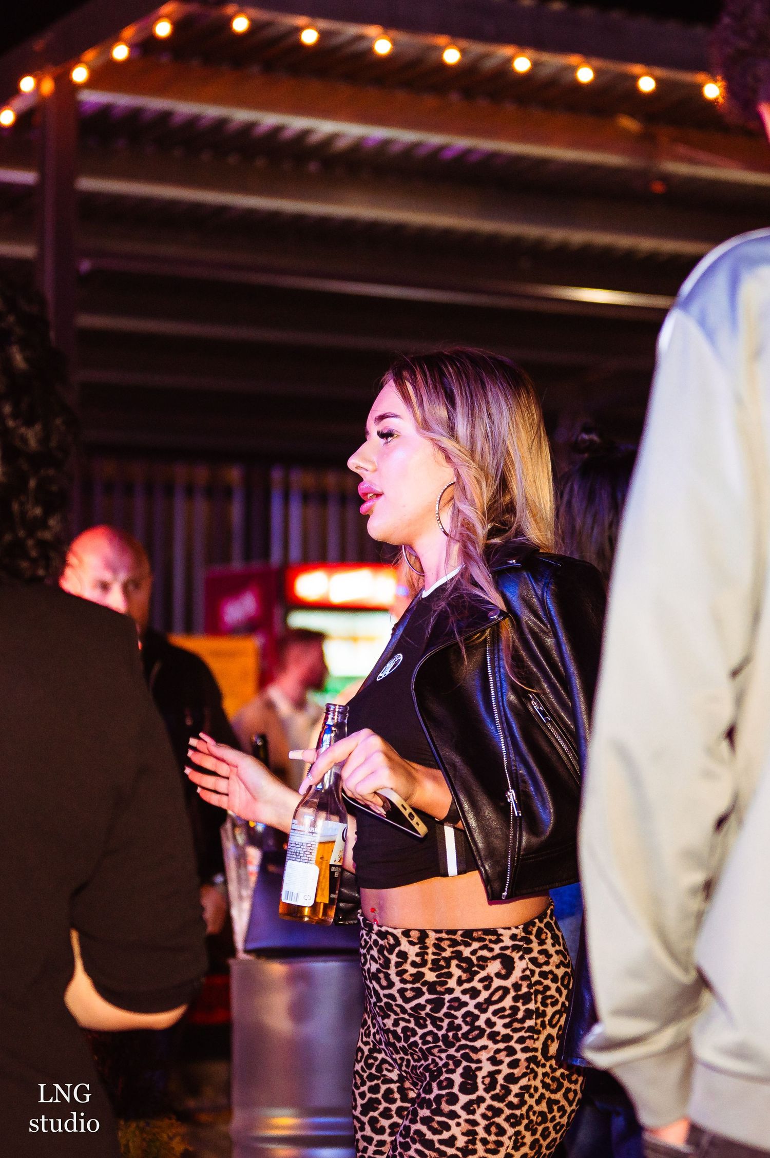 Party scene at night with someone in a leopard print skirt and leather jacket holding a beer bottle under string lights.
