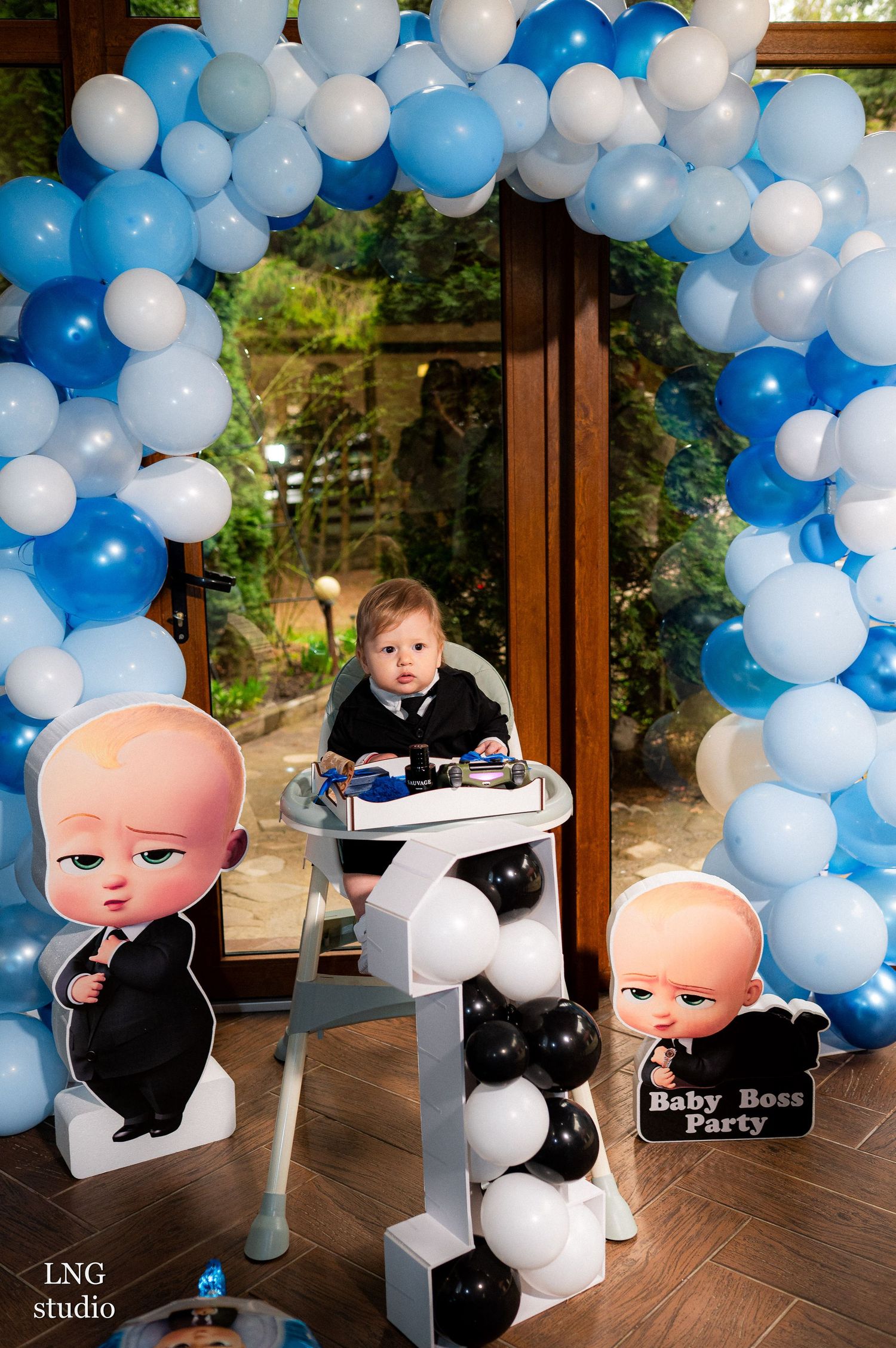 A first birthday party setup with blue and white balloon arch and Boss Baby themed cardboard cutout decorations.