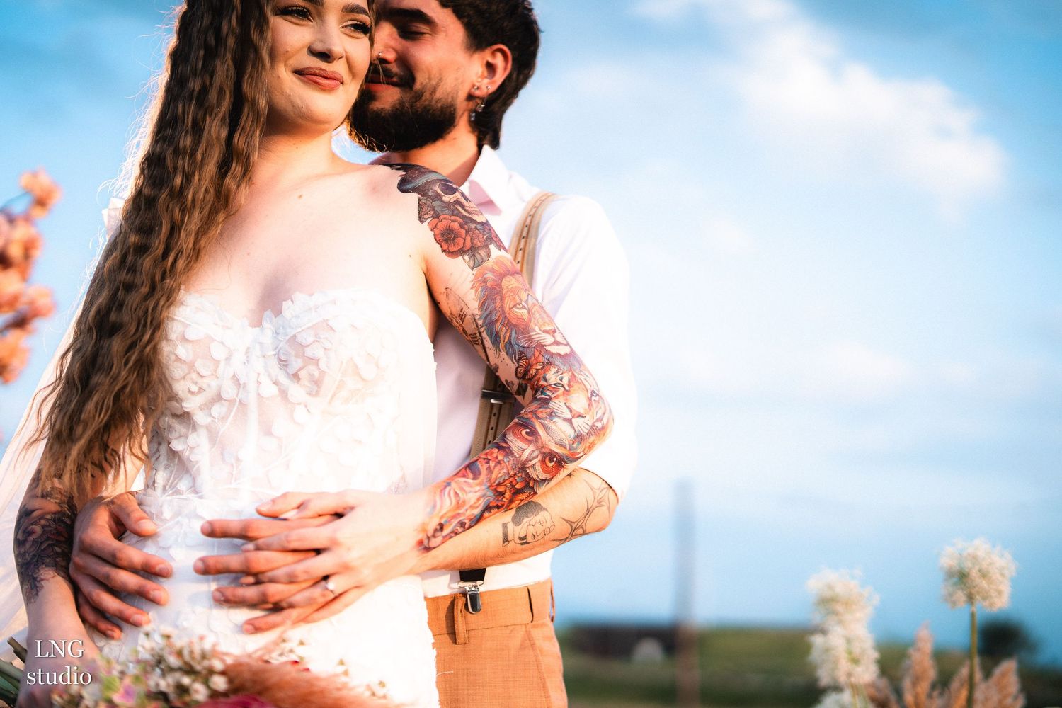 A romantic outdoor wedding photo captured at sunset with soft warm lighting and delicate white flowers in the background.