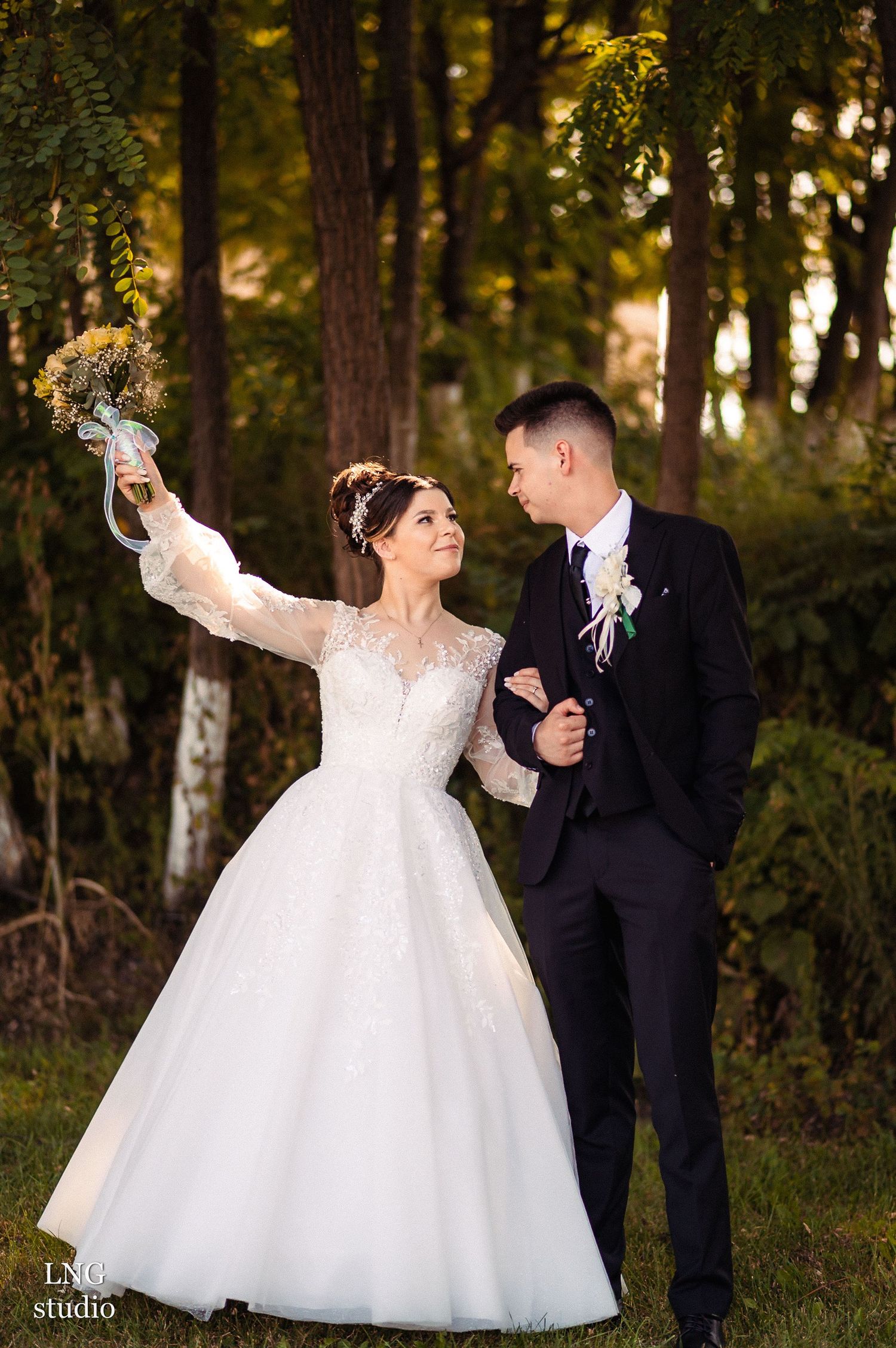 Bride in lace dress holds bouquet high while walking with groom through wooded setting.