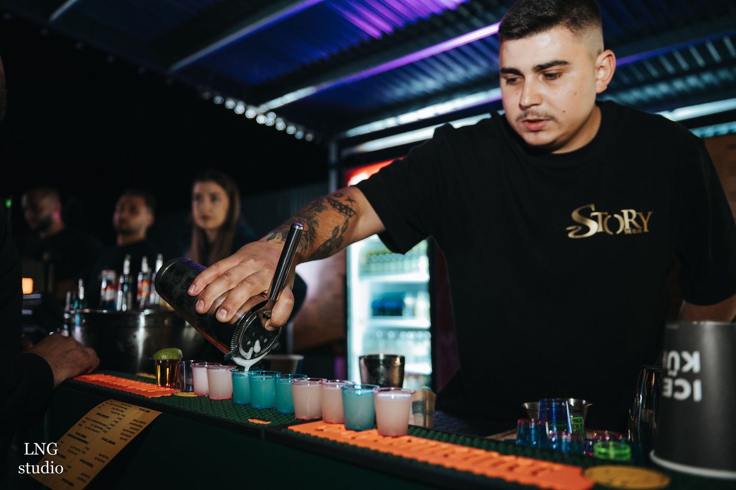 Dark moody bar scene with people sitting at counter while bartenders mix drinks.