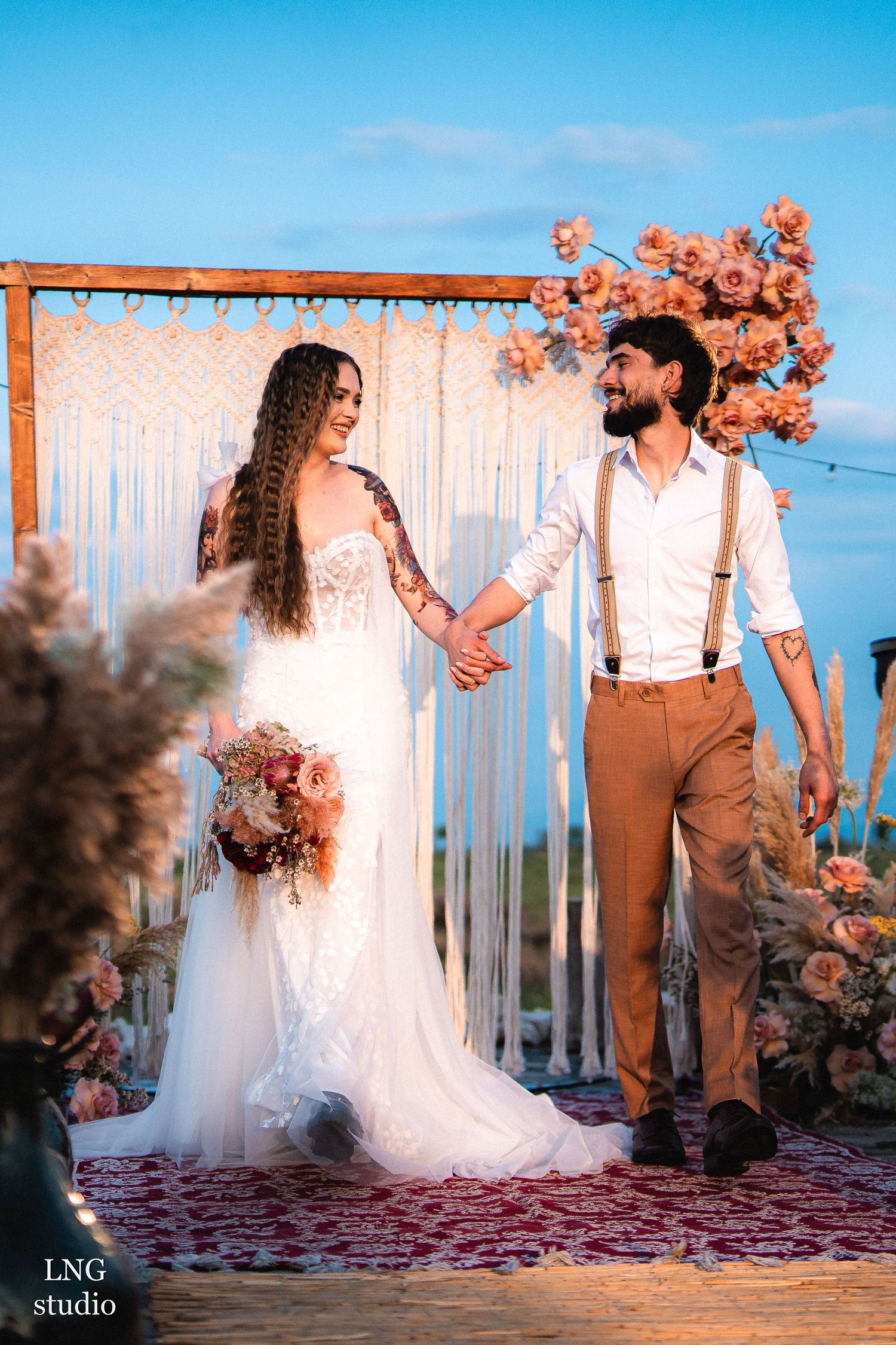 A romantic sunset photoshoot captures a couple in wedding attire standing by a rustic arch with peachy-pink floral decorations.