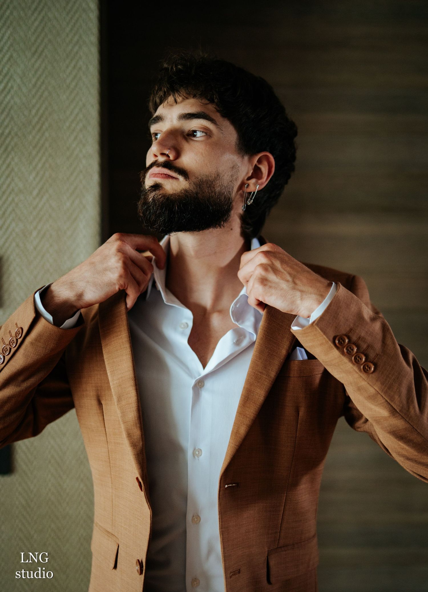 Portrait photo of a man adjusting his tan blazer collar while wearing a crisp white shirt in moody lighting.
