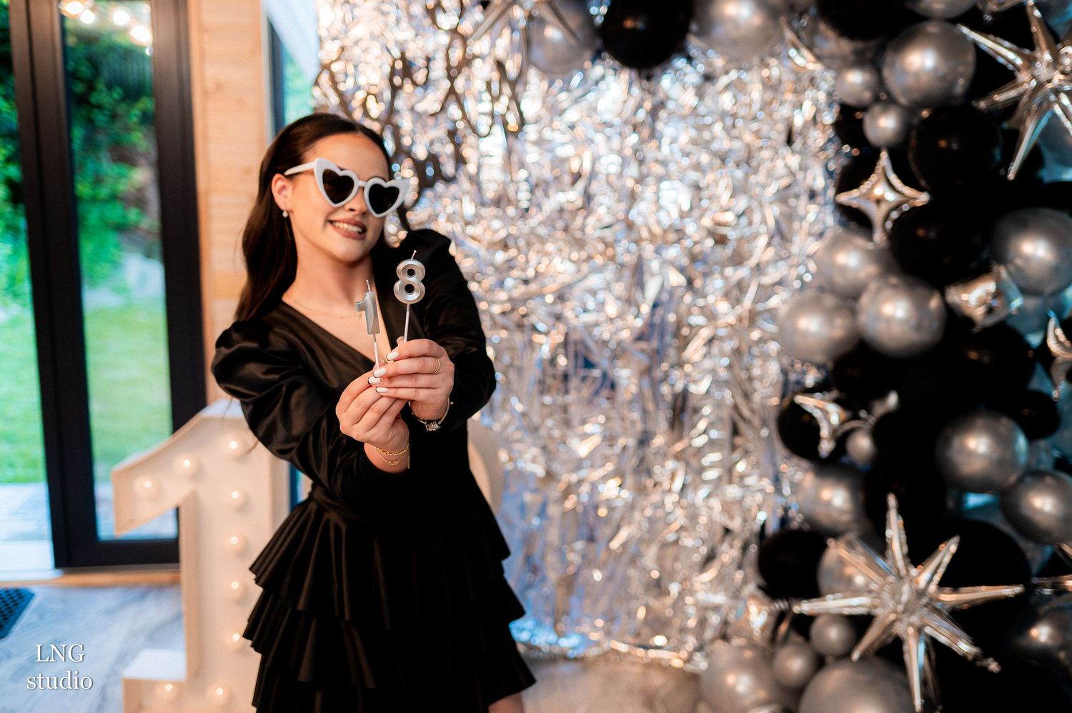 Someone in a black velvet dress and sunglasses holds a champagne glass against a festive silver and black holiday backdrop.