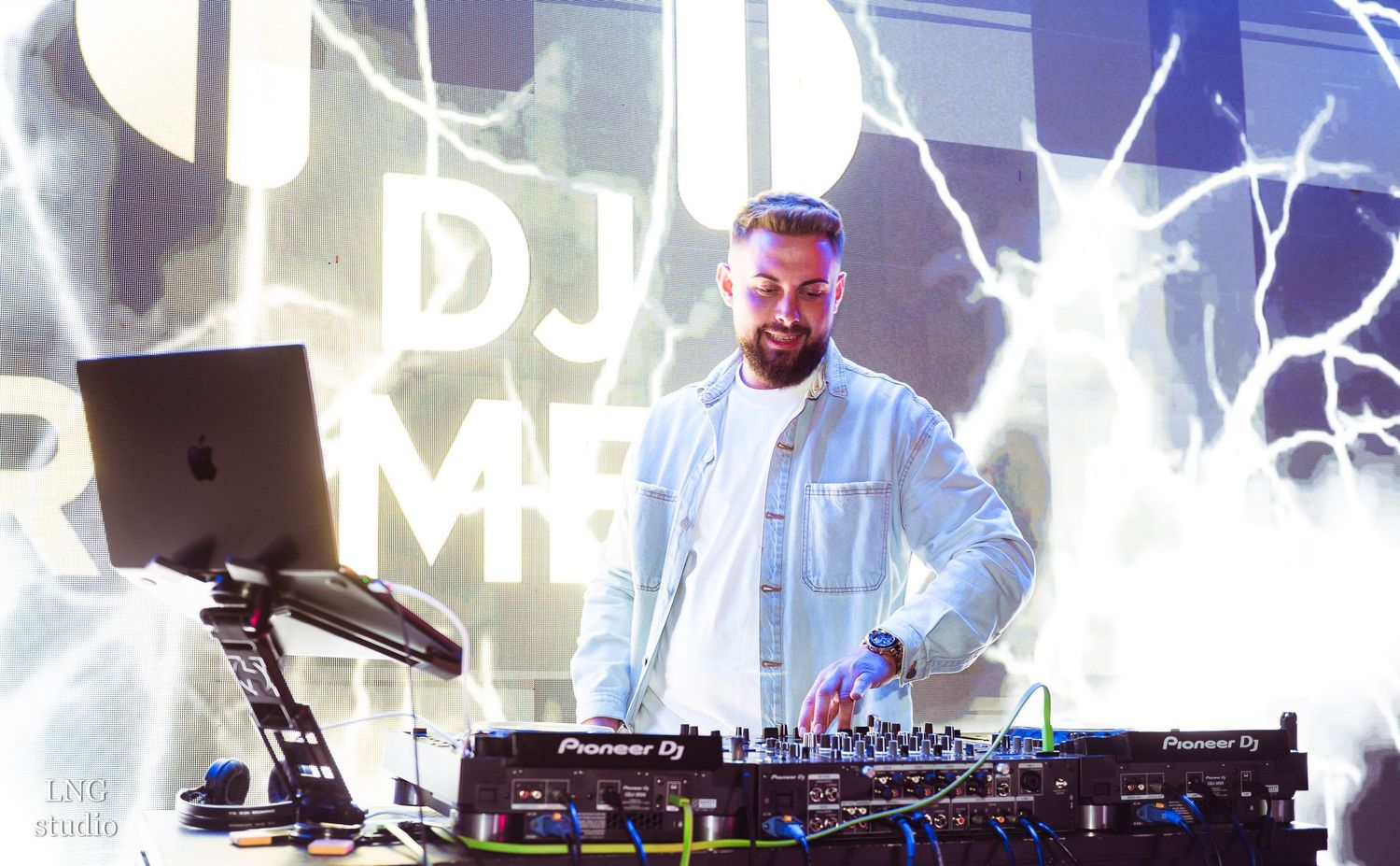 DJ performing with laptop on stage with bright lightning effect visuals in background.