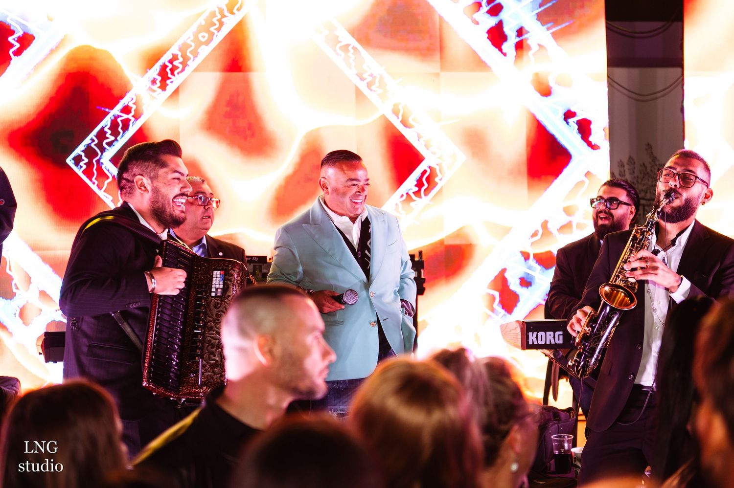 Musicians perform with accordion and brass instruments against geometric red backdrop on stage.