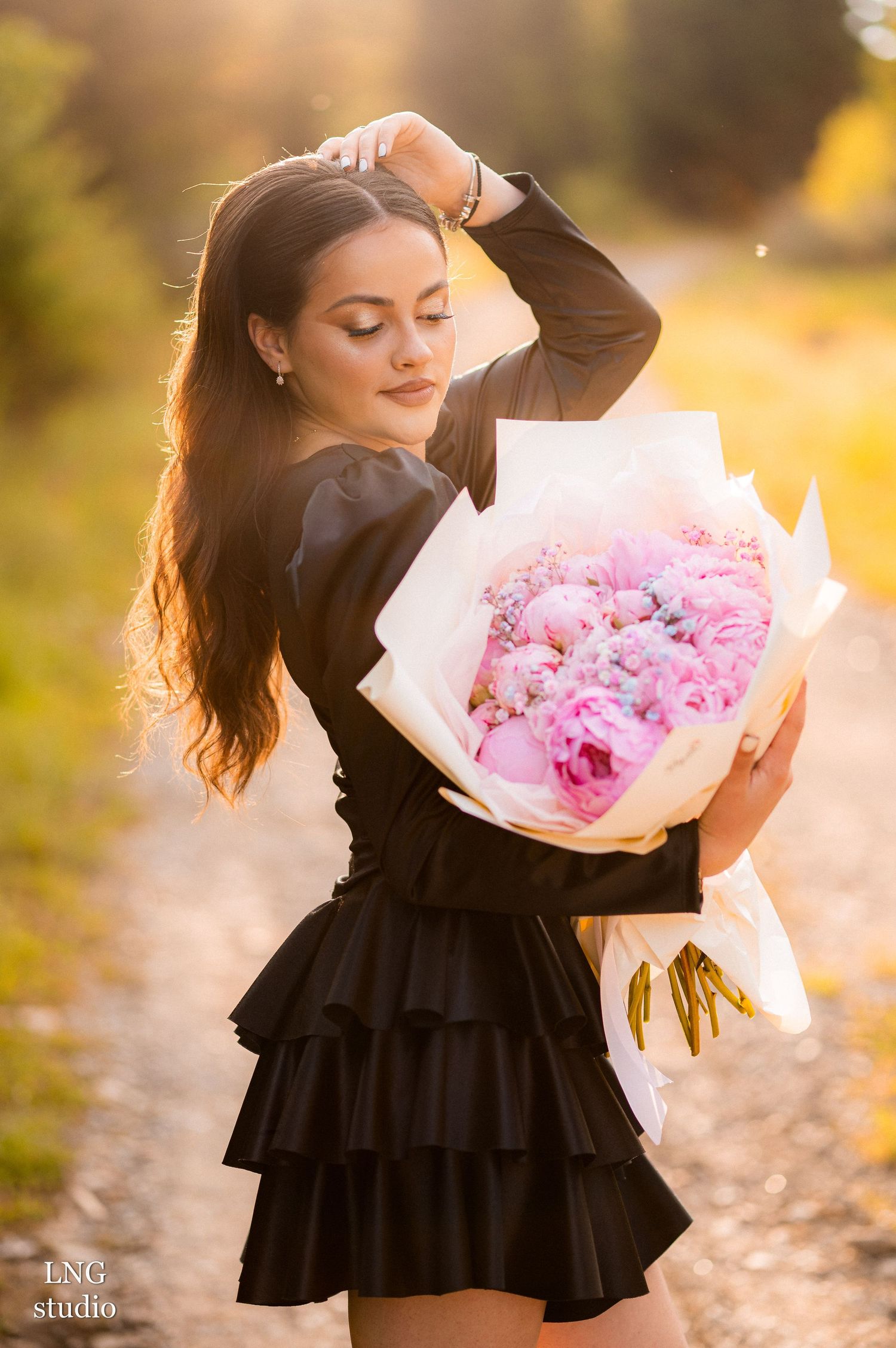 A black dress adorned figure holds a lush bouquet of pink peonies in golden sunset lighting outdoors.