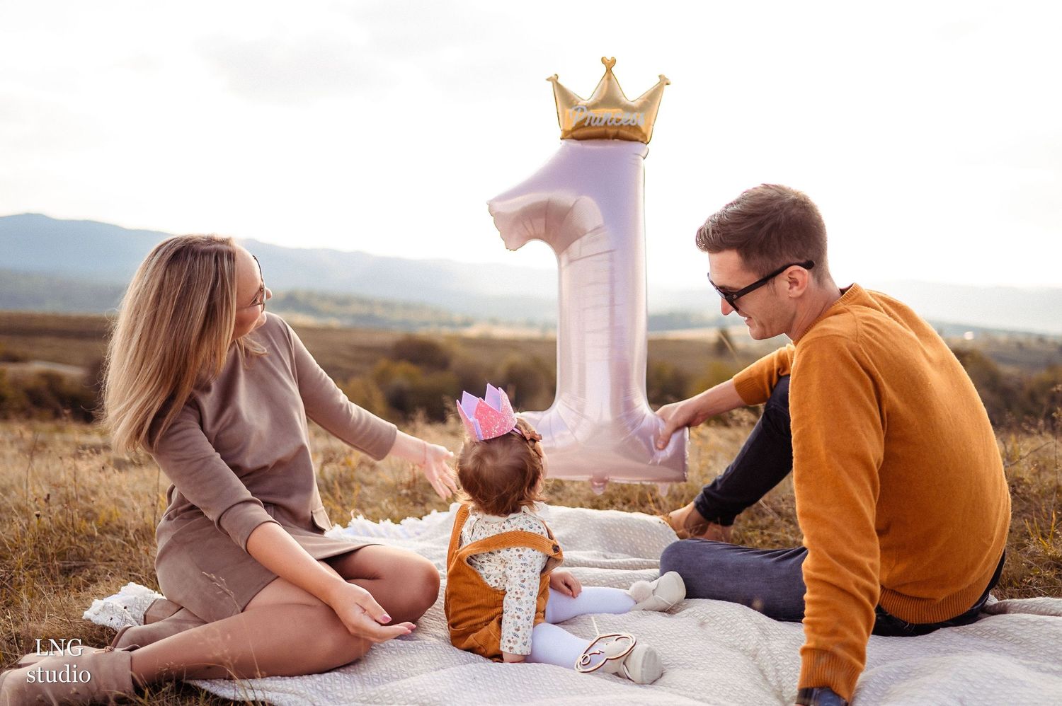 Family celebrates first birthday with large number one decoration and crown outdoors.