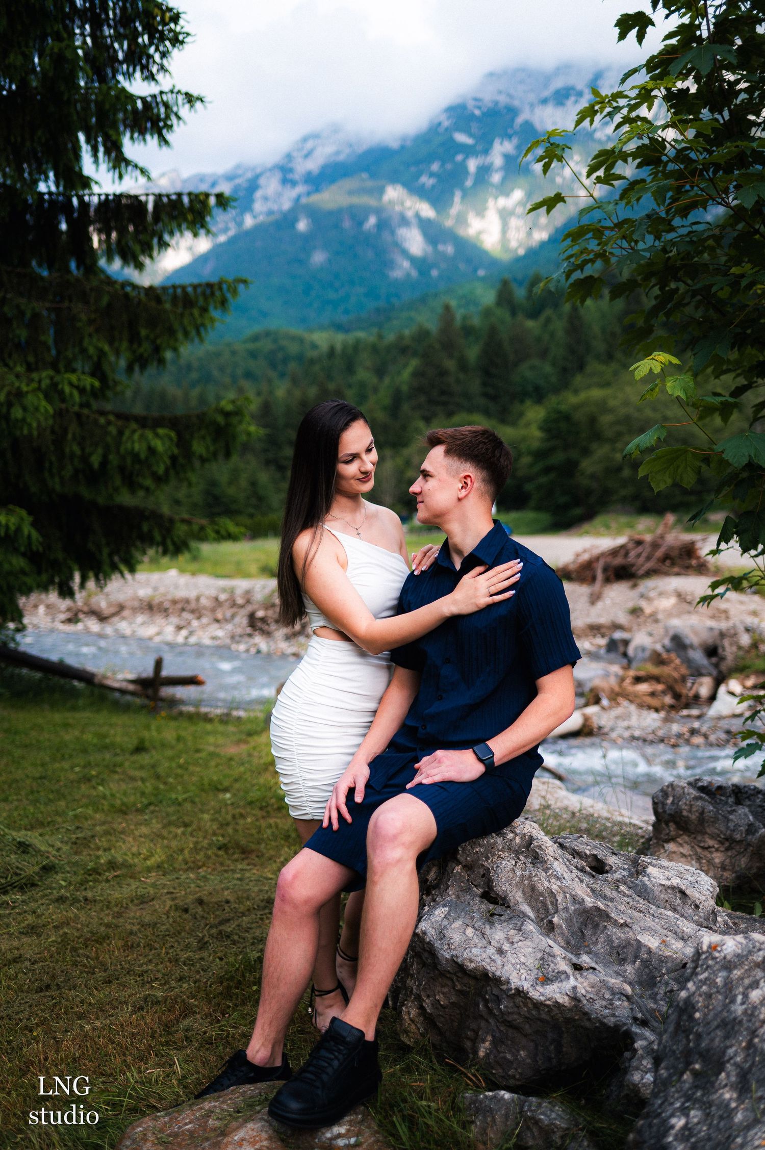 A couple sits together on rocks by a mountain stream during a photo session.