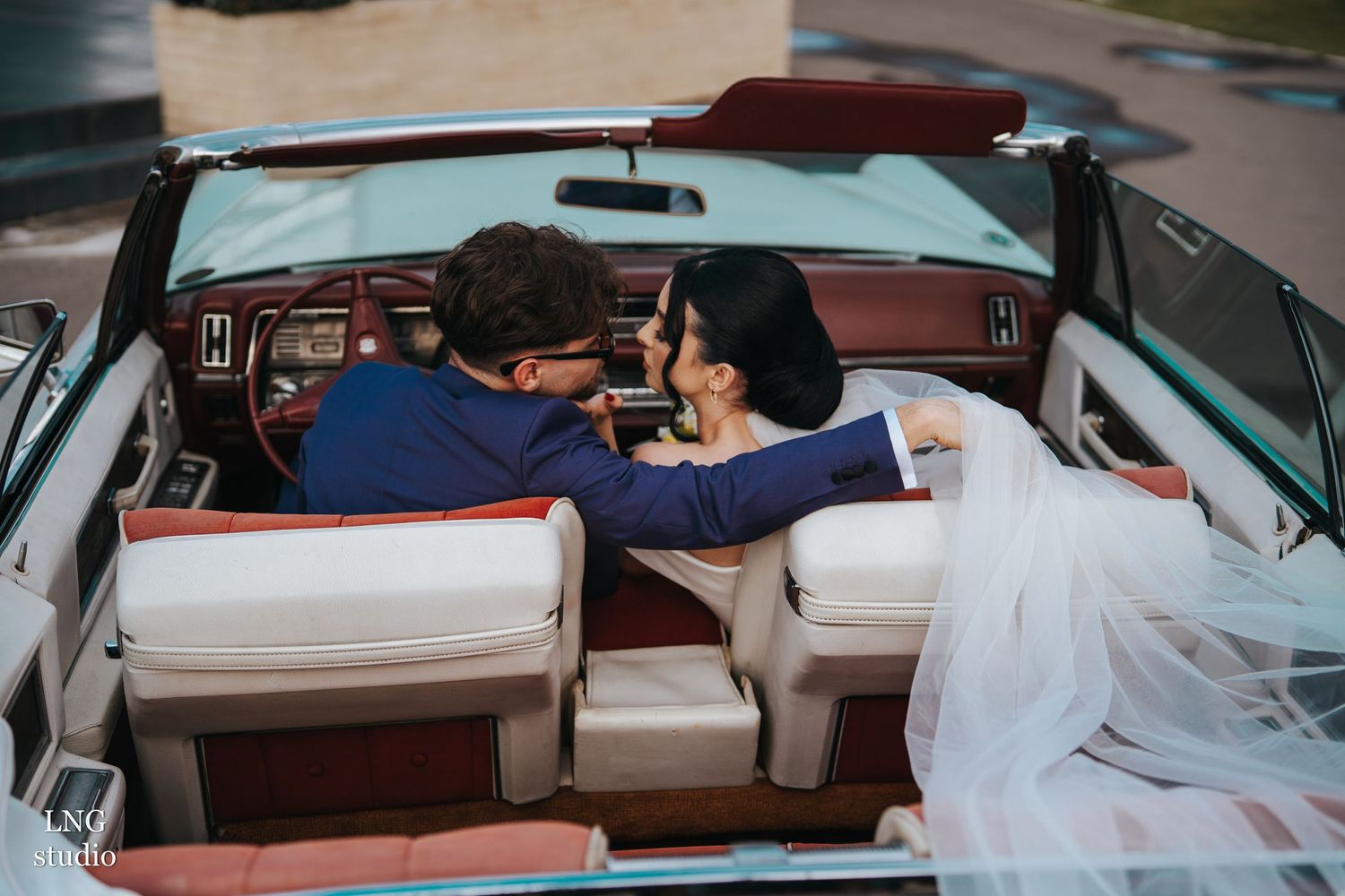 A couple sits close together in a vintage convertible car while wearing wedding attire.