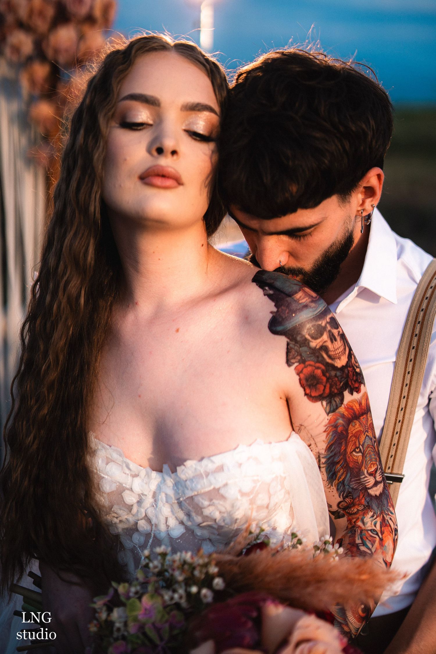 Romantic portrait of a couple in bridal attire with tattoos in warm sunset lighting outdoors.