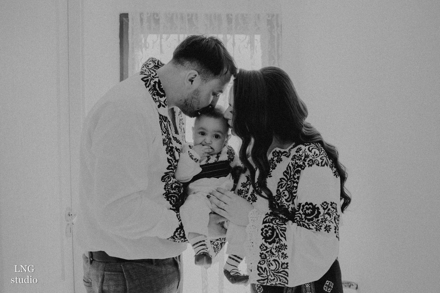 Black and white portrait of family sharing tender moment in traditional embroidered clothing.