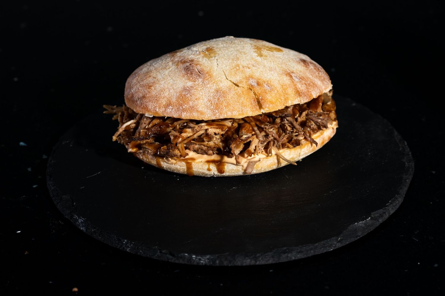 A pulled pork sandwich on artisanal bread served on a black slate plate.