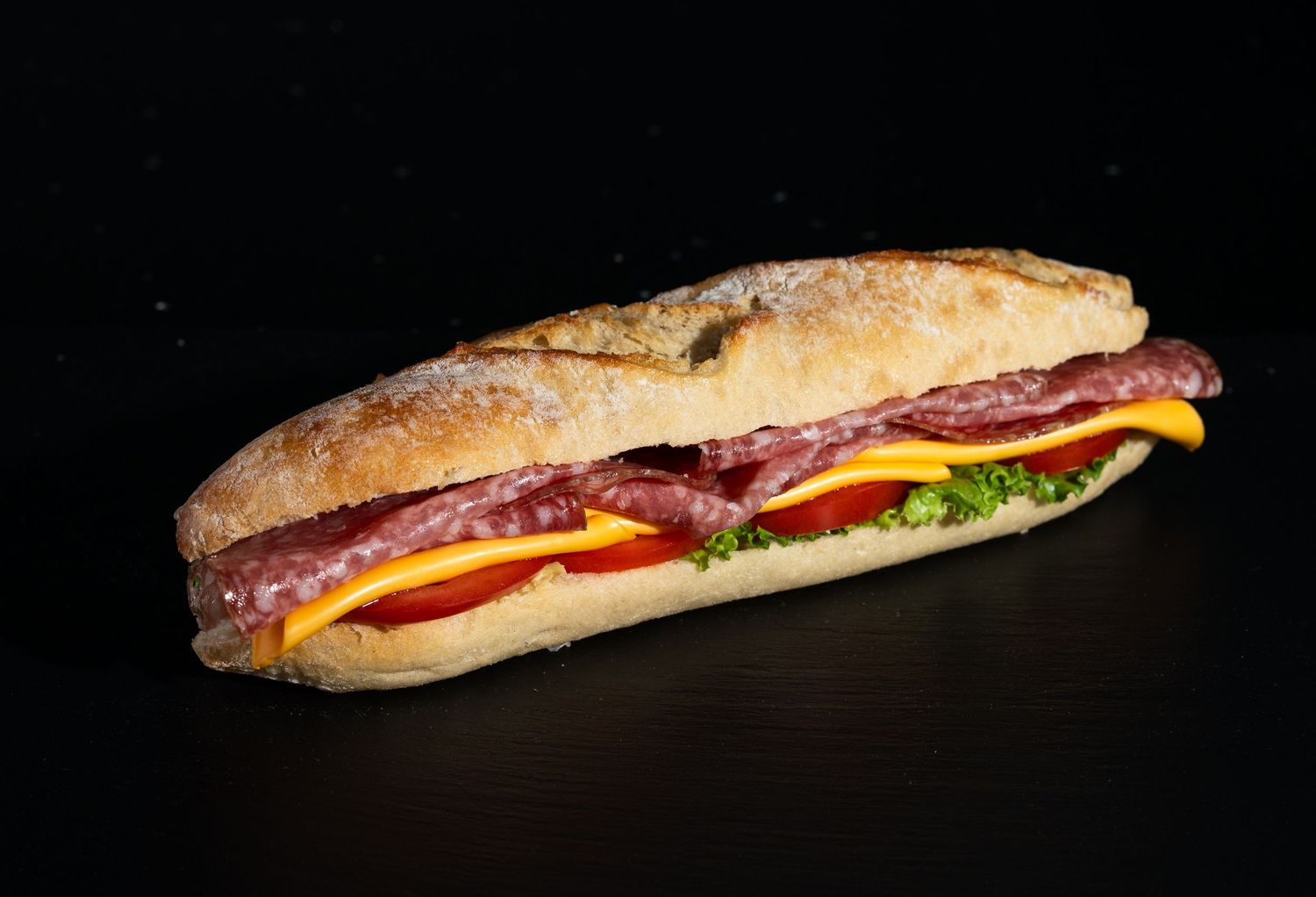 Fresh submarine sandwich with meat, cheese, lettuce and tomatoes on crusty bread photographed against black background.
