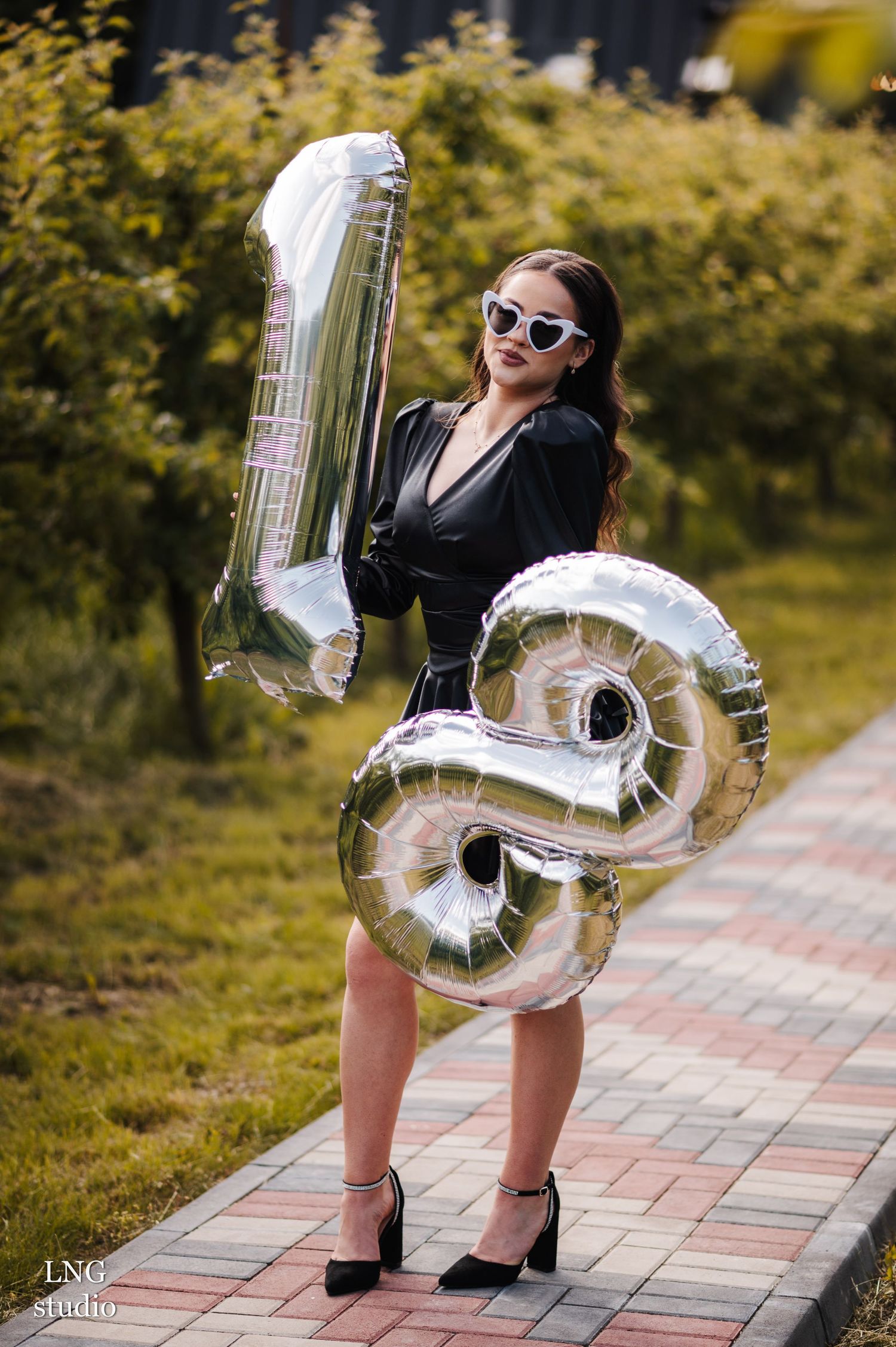 Stylish silver number balloons spelling '18' held outdoors on a brick pathway with green foliage in background.