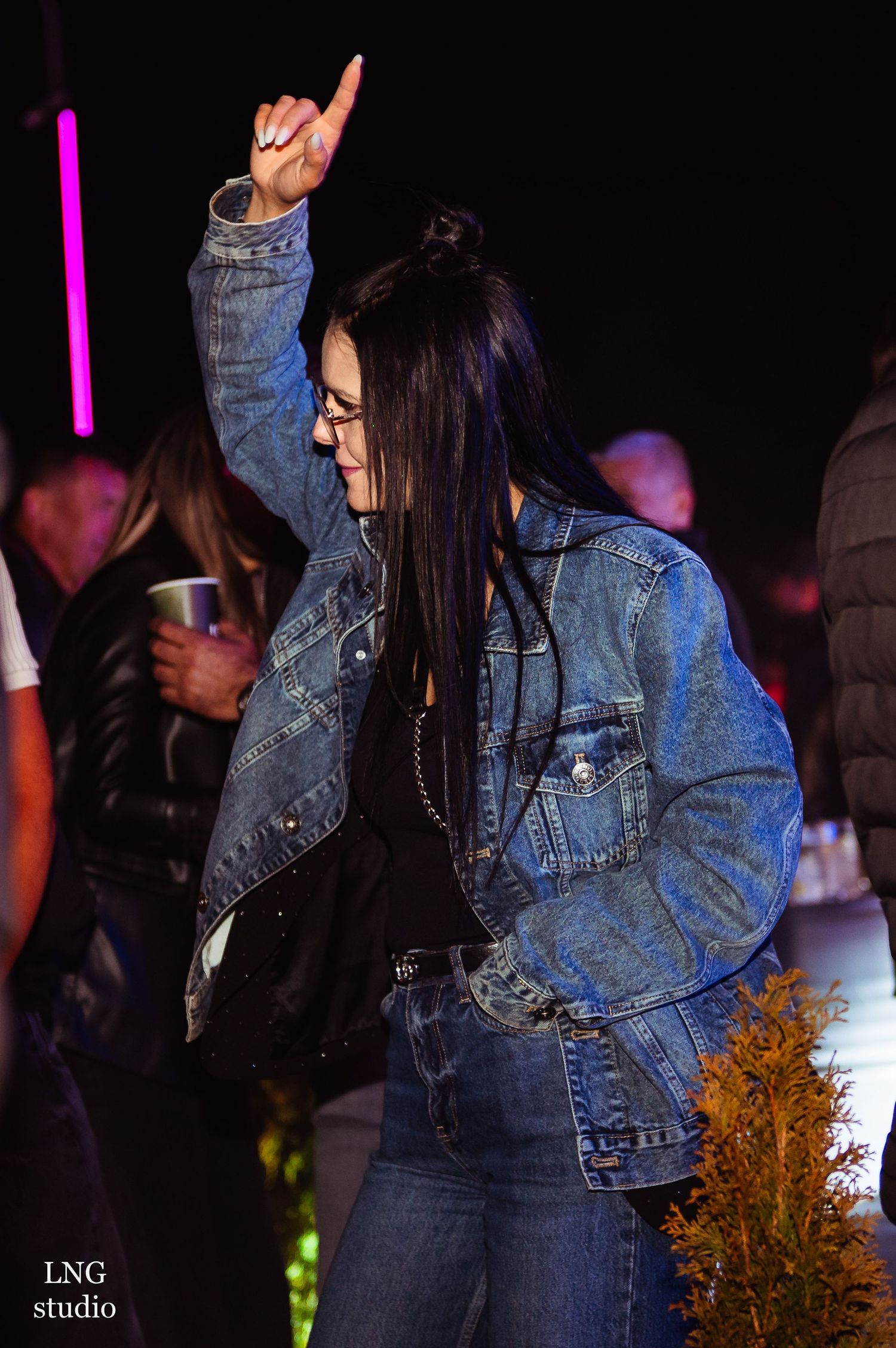 Person in denim jacket dancing with raised arm at a dark nightclub setting illuminated by purple lighting.