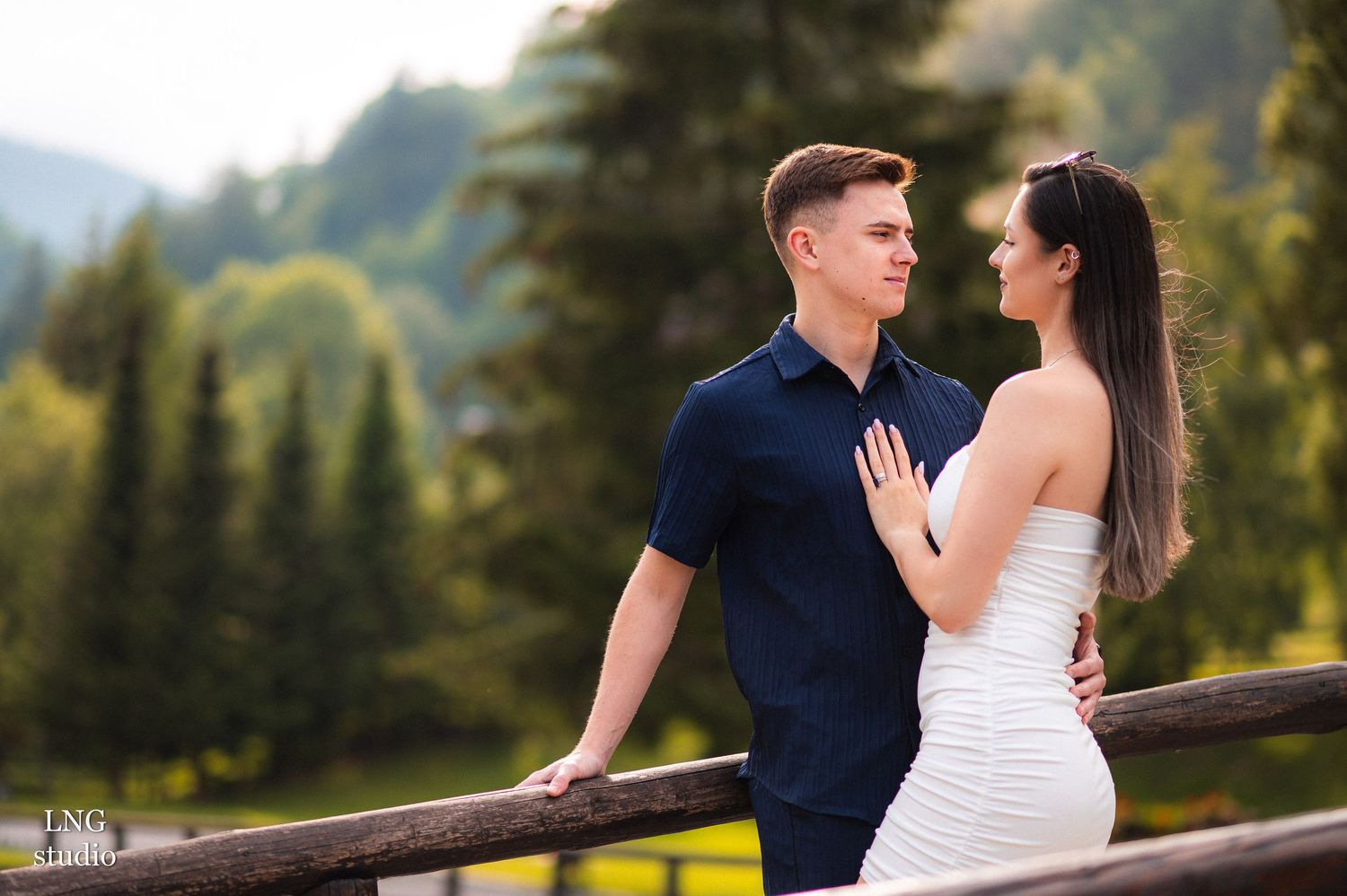 Romantic couple poses near rustic wooden fence with mountain scenery.