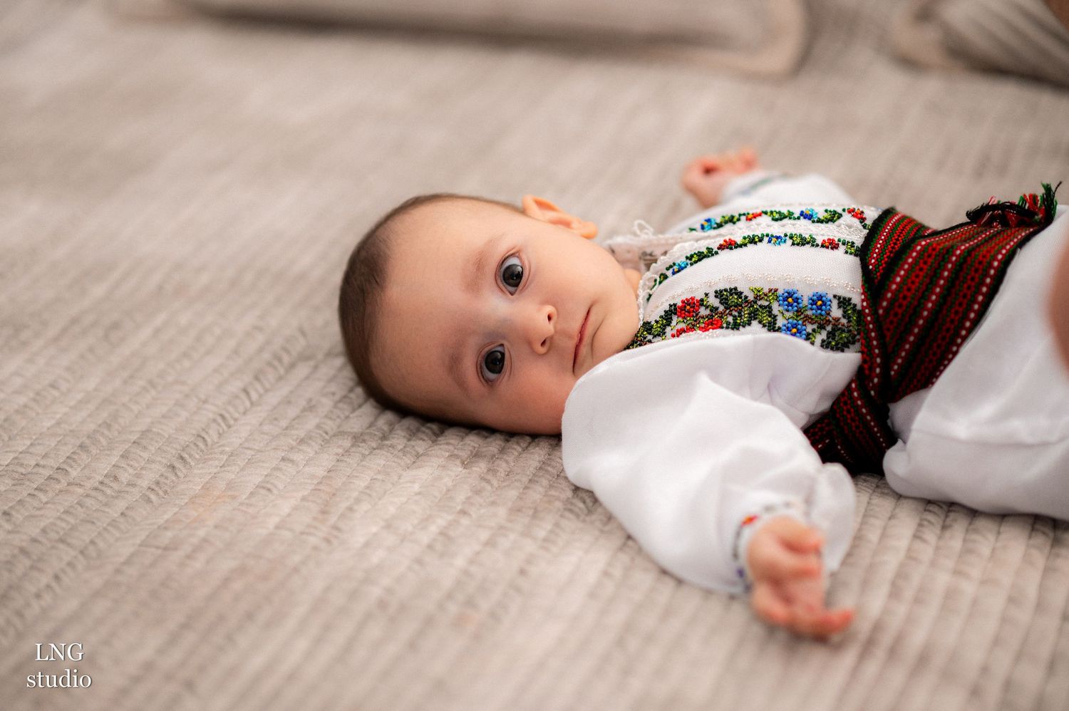 Baby lying on soft gray textured surface in traditional embroidered clothing.