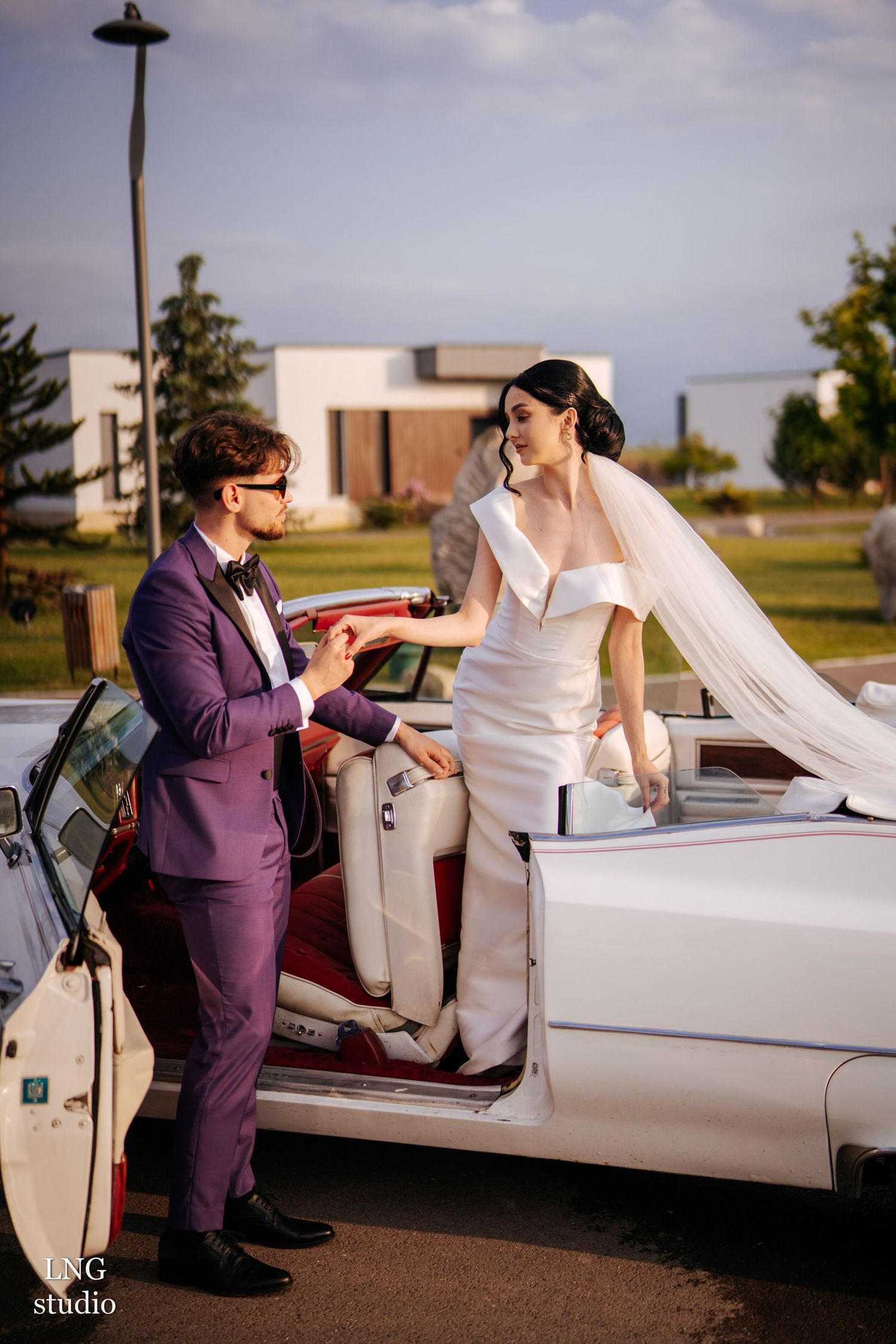 A couple in wedding attire stand beside a classic silver car, with one in a purple suit helping the other exit the vehicle.