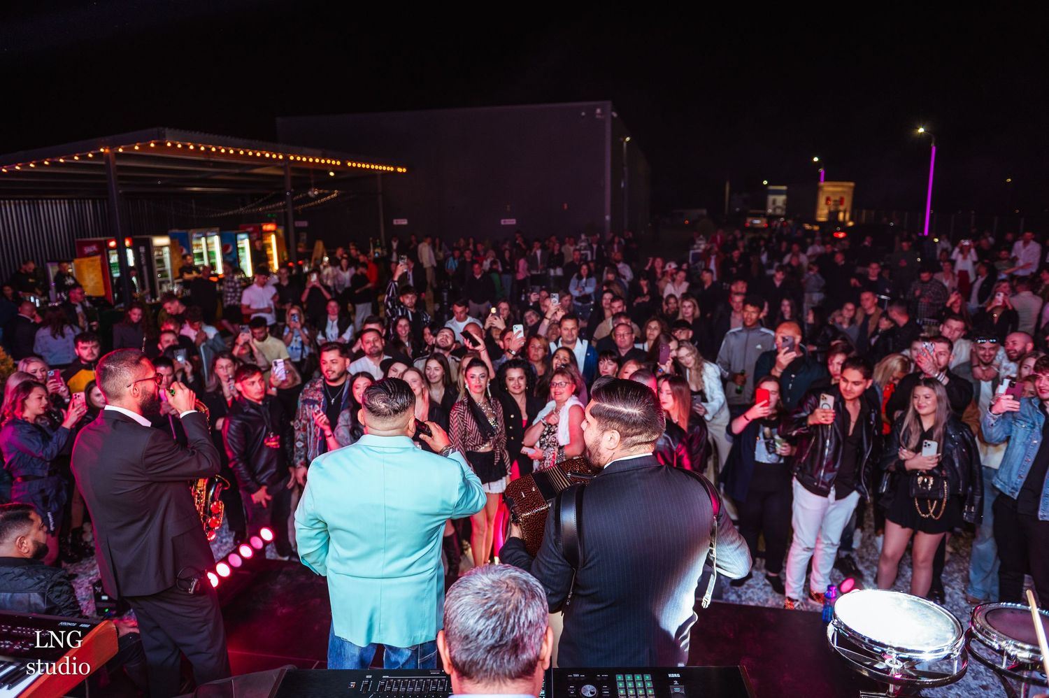 Large crowd gathers at outdoor concert venue with band performing on stage under colored lights.