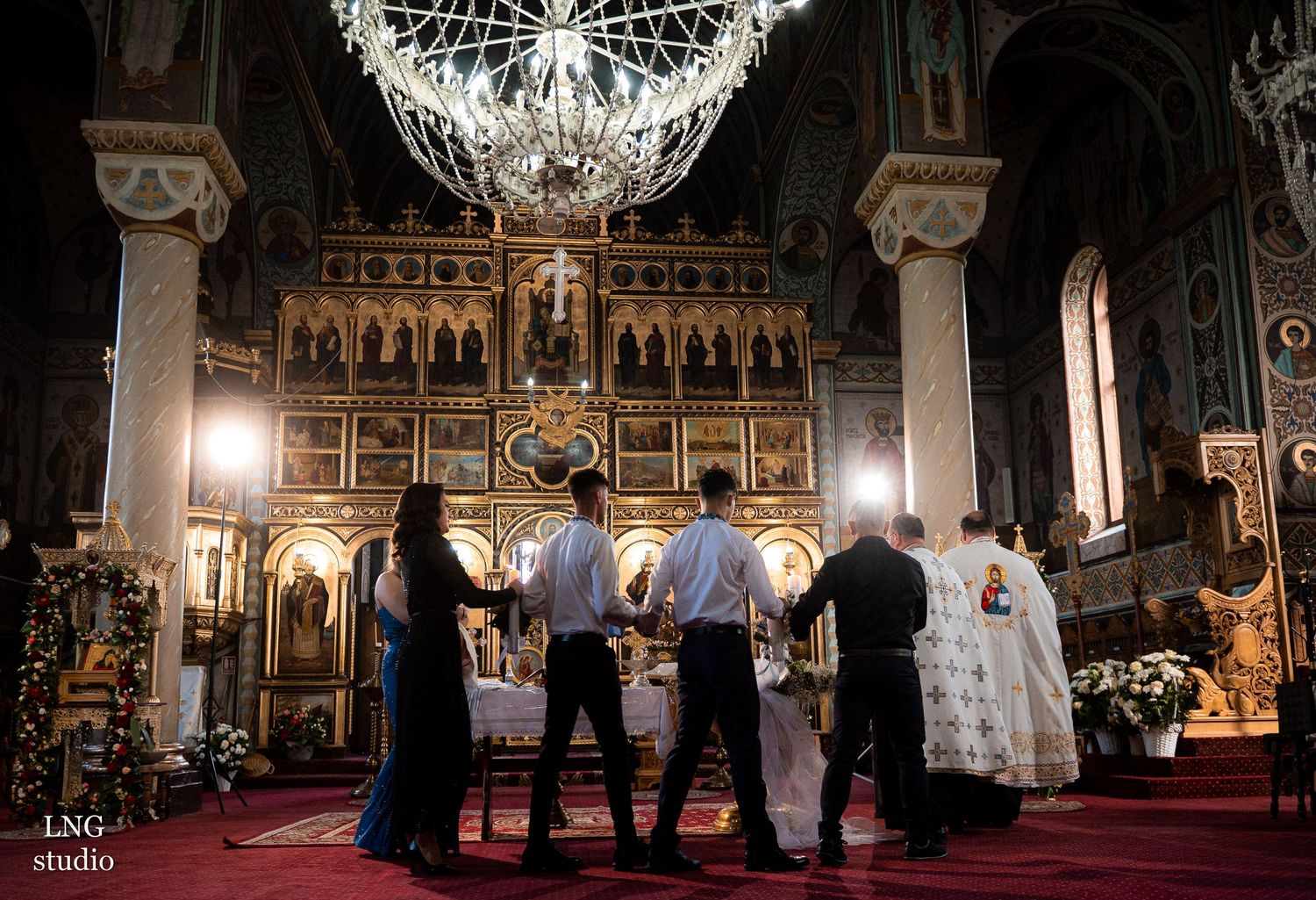 Orthodox priests conduct a religious ceremony in an ornate church with a grand chandelier and iconostasis.