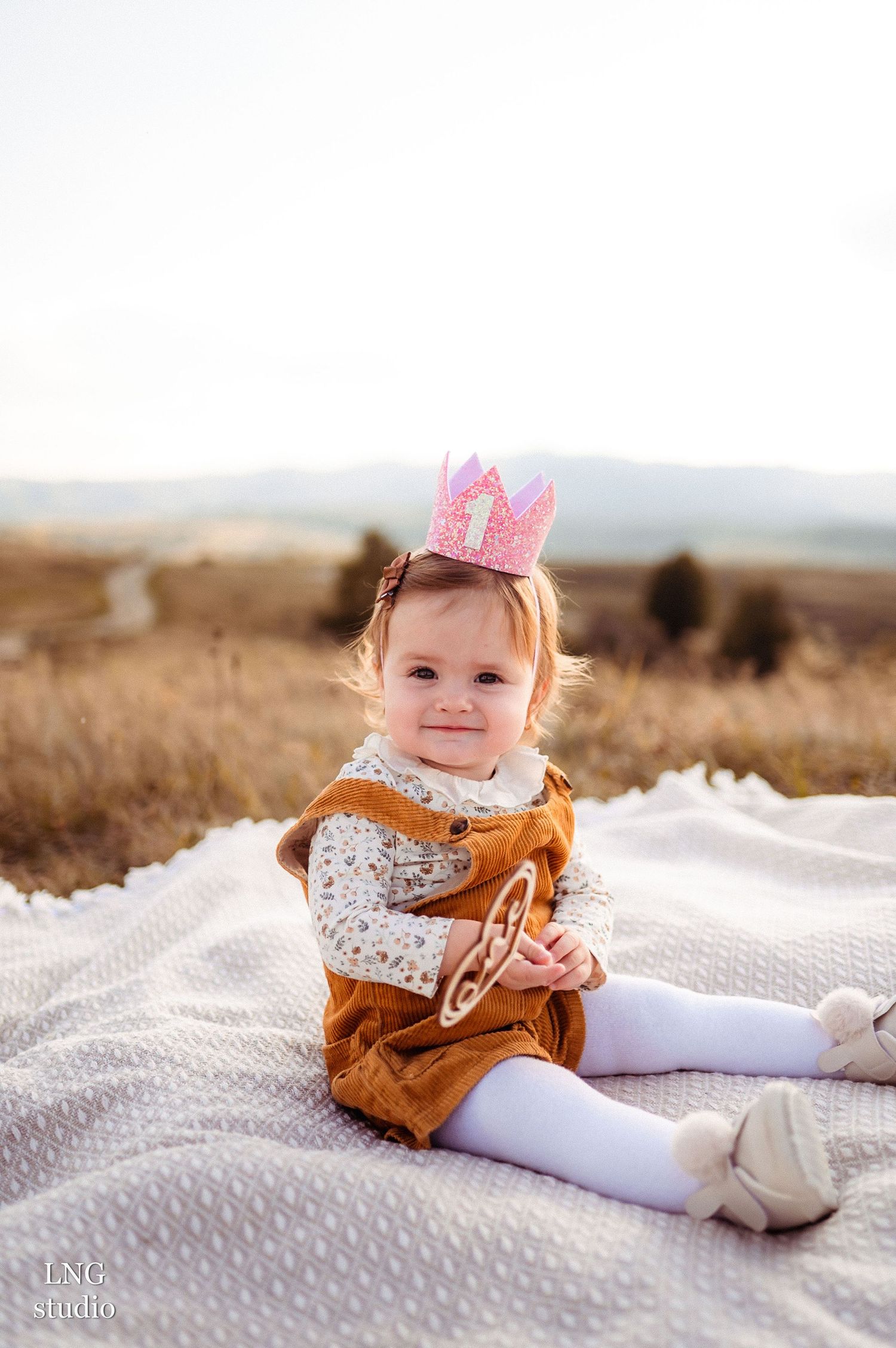 Toddler wearing orange corduroy dress and pink crown sits on white blanket in outdoor field setting.