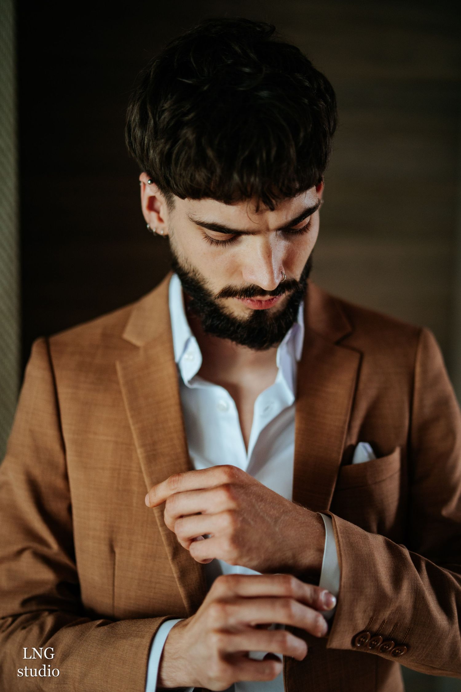 A bearded man in a brown suit and white shirt adjusts his cuff in a moody portrait lighting setup.