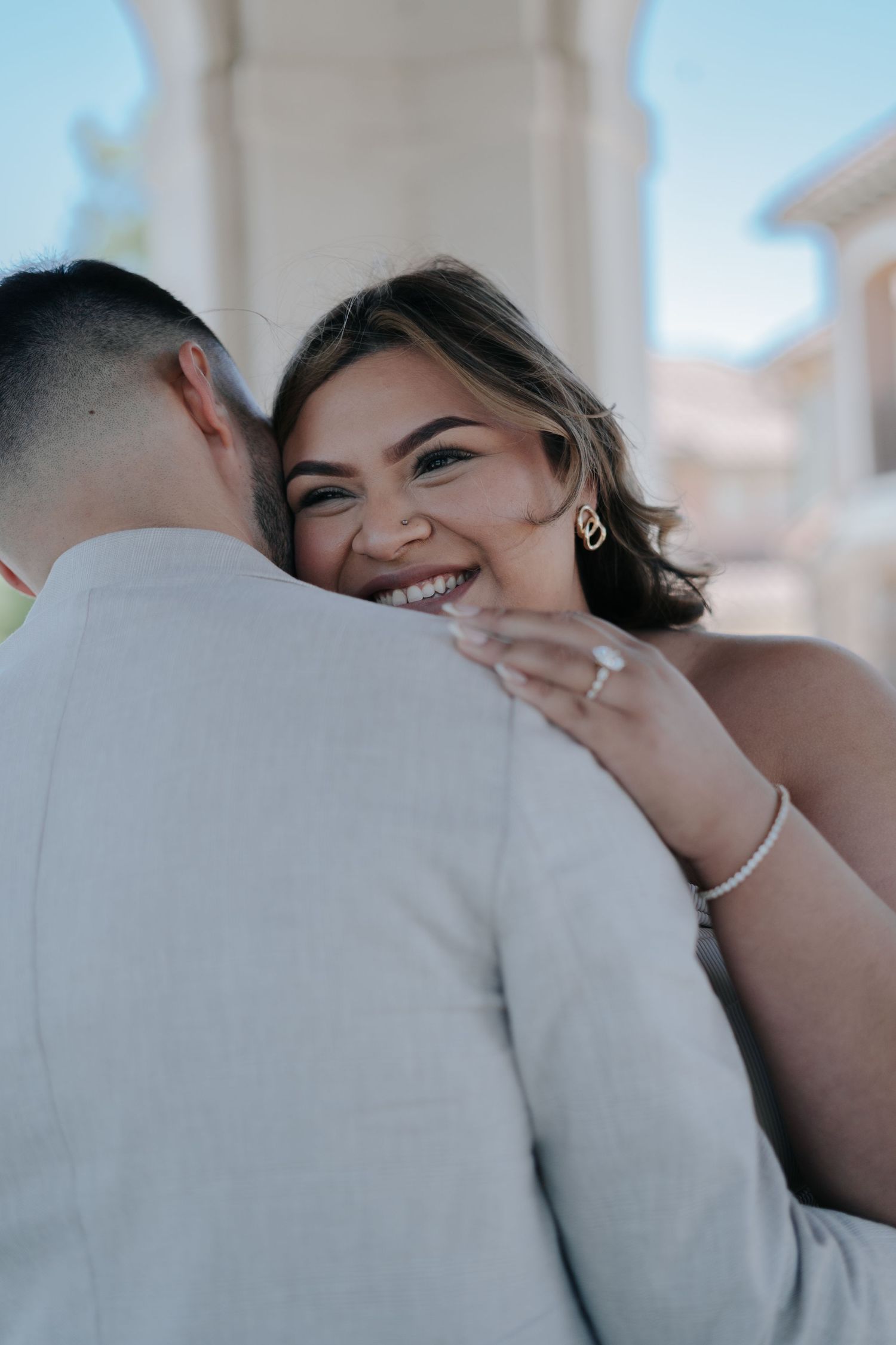 Smiling bride in white dress embraces groom in light gray suit during outdoor wedding ceremony photo.