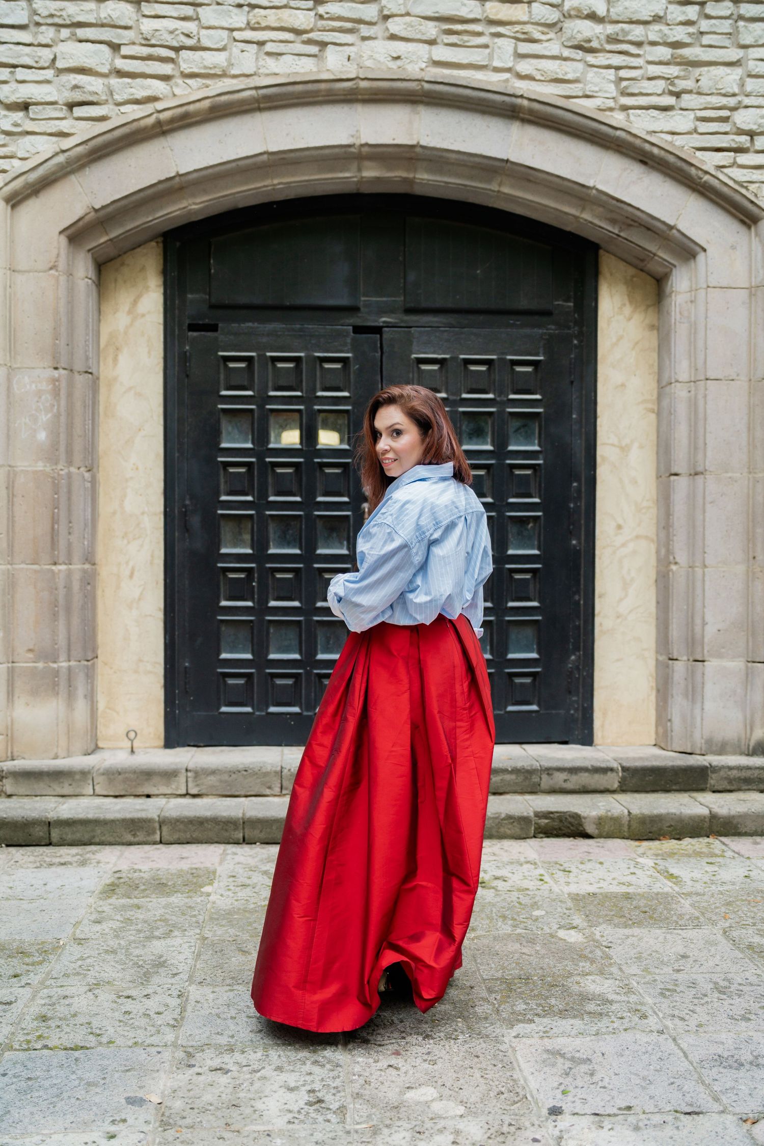 Woman in red satin maxi skirt and blue blouse poses before ornate stone archway with black wooden door.