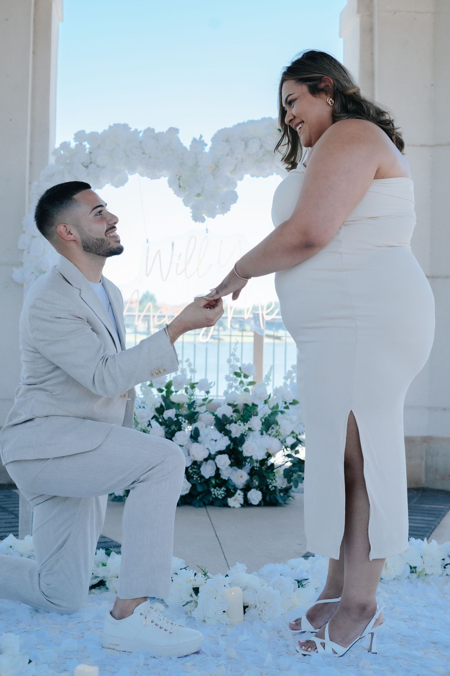 Man kneeling on one knee proposing to woman in white dress surrounded by white flowers and heart backdrop.