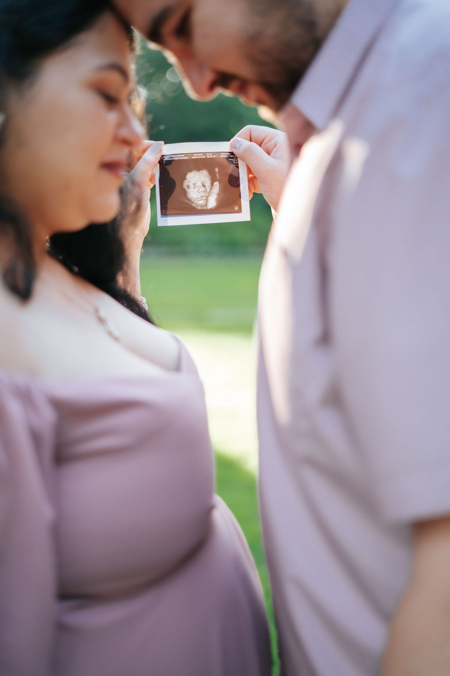 Pregnant couple holding ultrasound photo outdoors, woman in pink dress, soft focus green background.