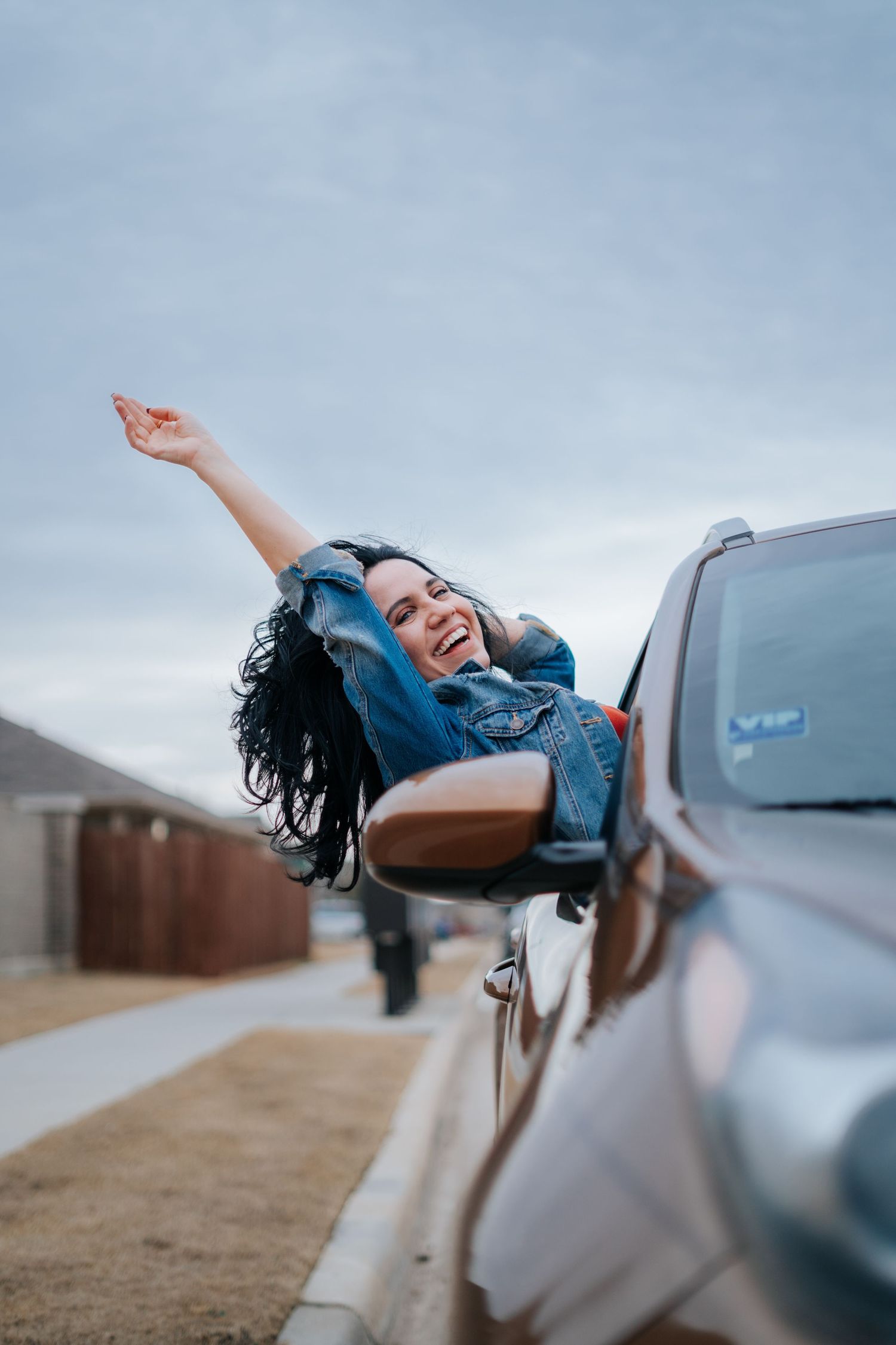 A joyful woman in a denim jacket leans out of a car window, laughing and raising her arm in celebration.