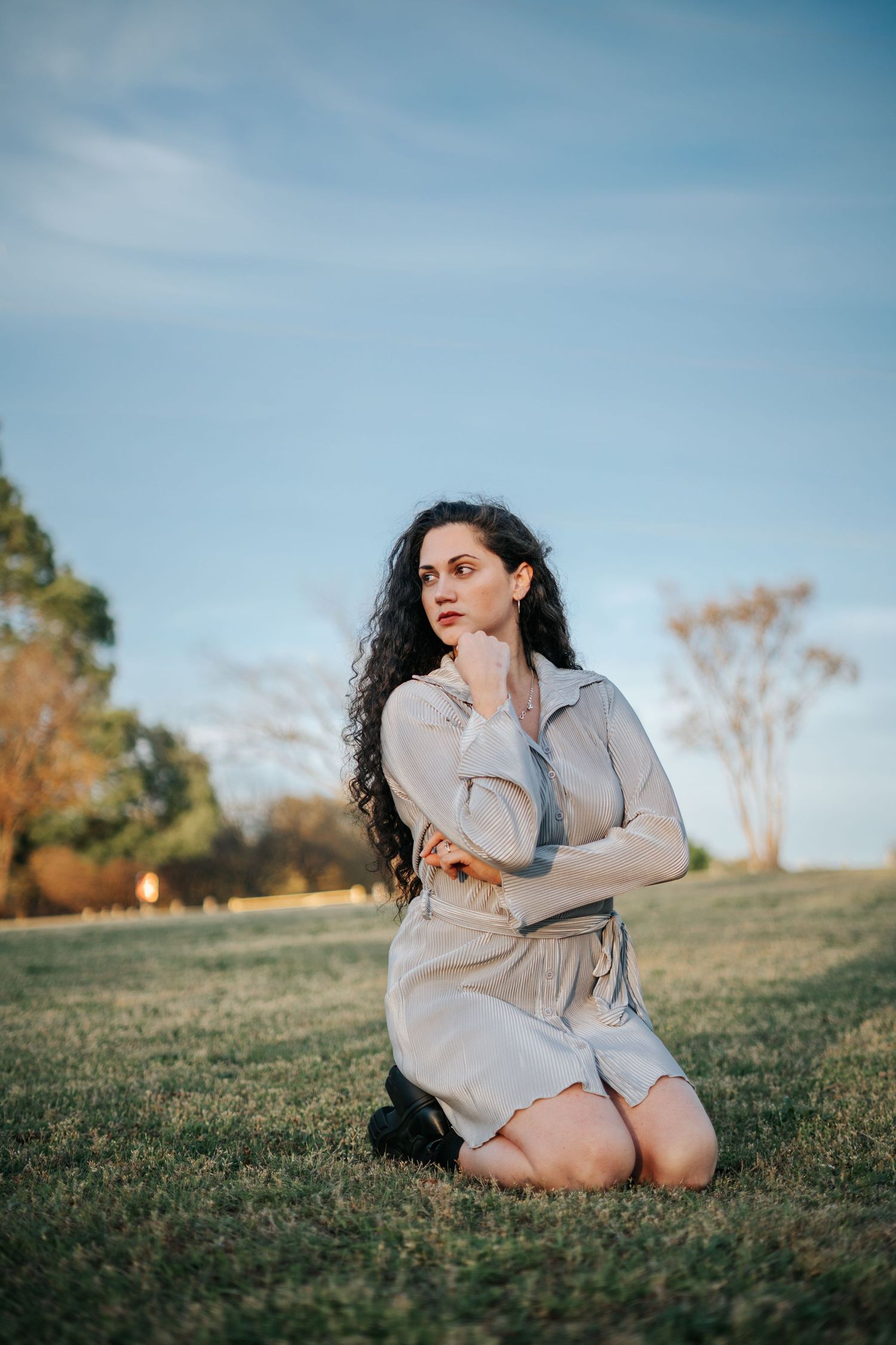 Woman in beige dress kneeling on grass in a park, gazing thoughtfully into the distance under a blue sky.