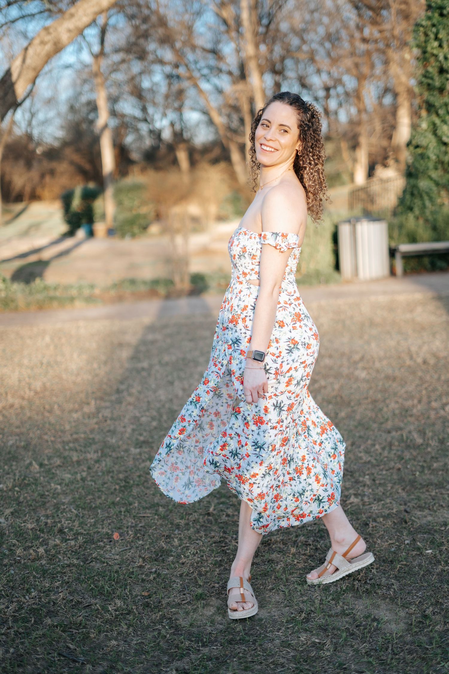 Woman in floral off shoulder midi dress smiling in a sunlit park, curly hair flowing, wearing sandals.