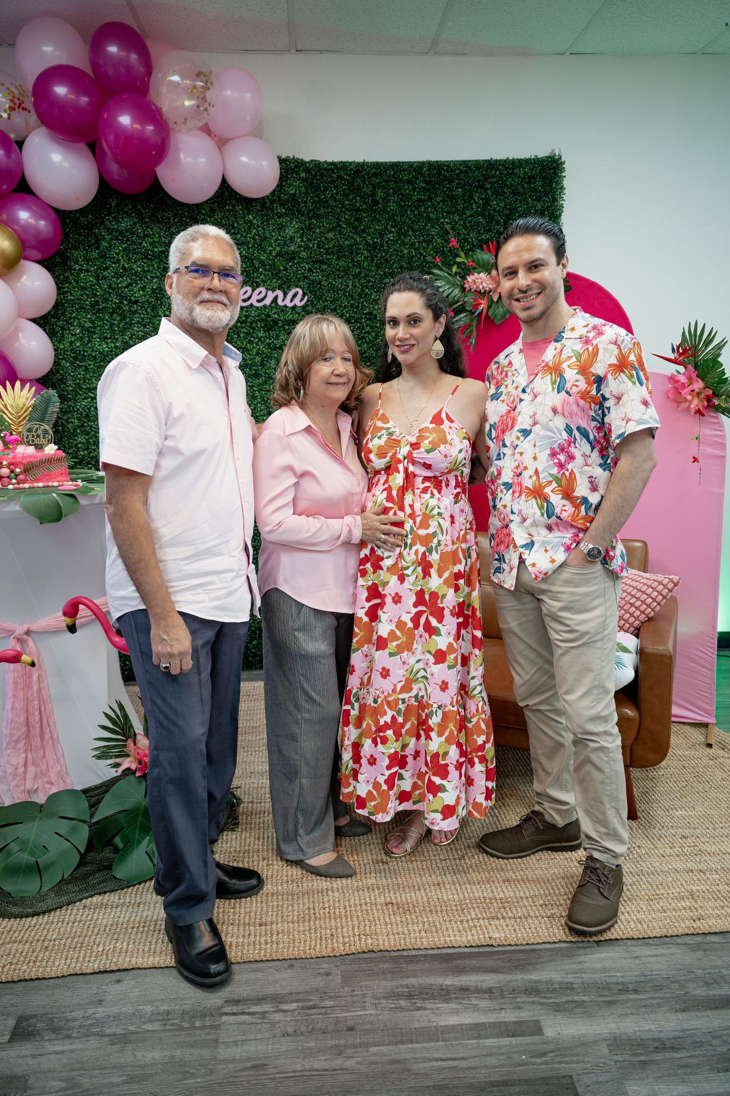 Four people posing together at a tropical flamingo themed party with pink balloons and a green hedge backdrop.