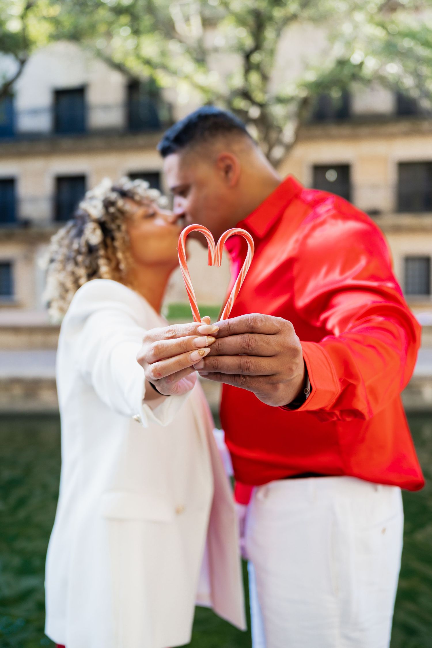 Couple kissing while holding two candy canes forming a heart shape outdoors, wearing red and white outfits.
