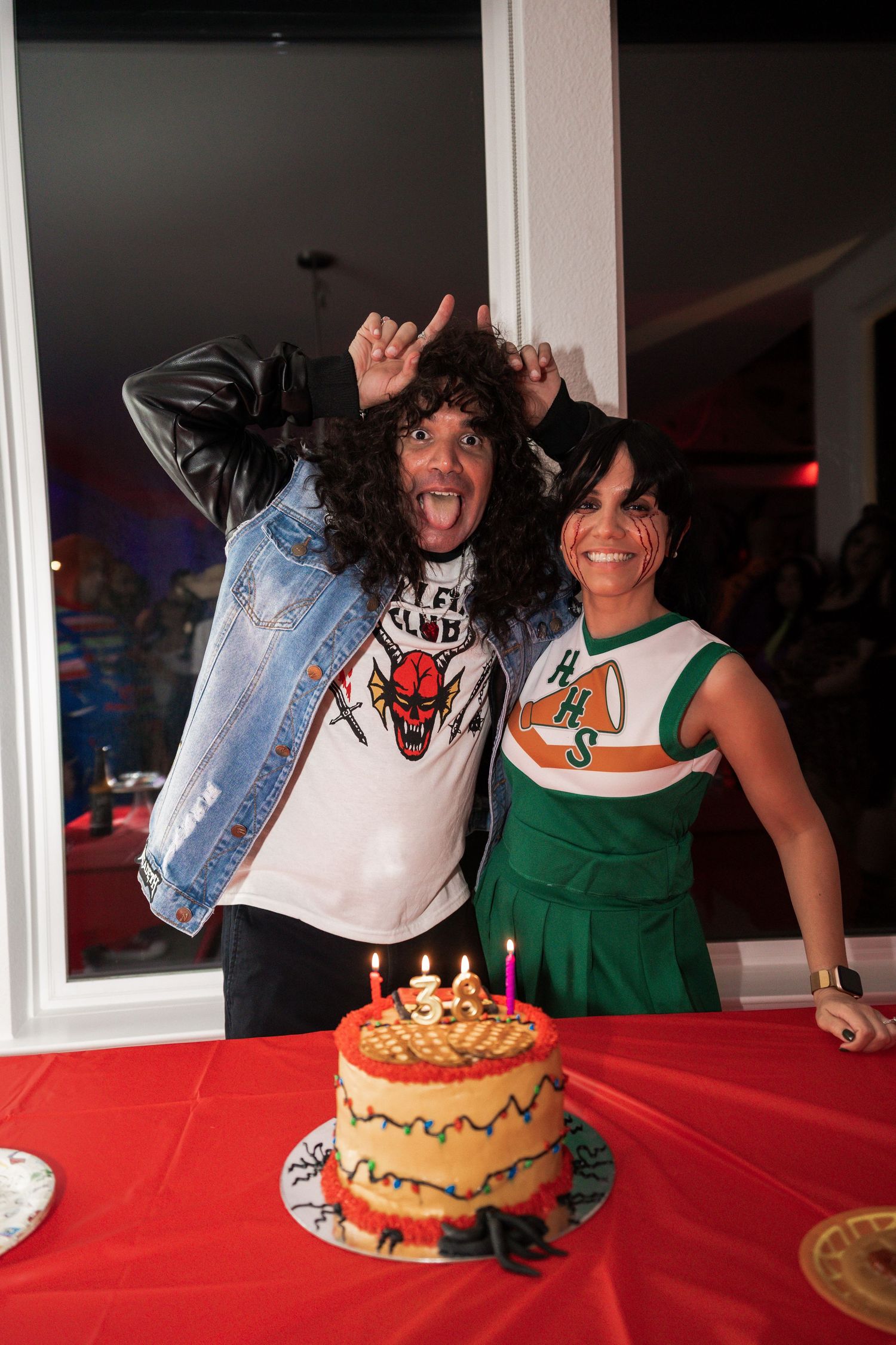 Two people in Halloween costumes pose with a decorated birthday cake with candles on a red table.